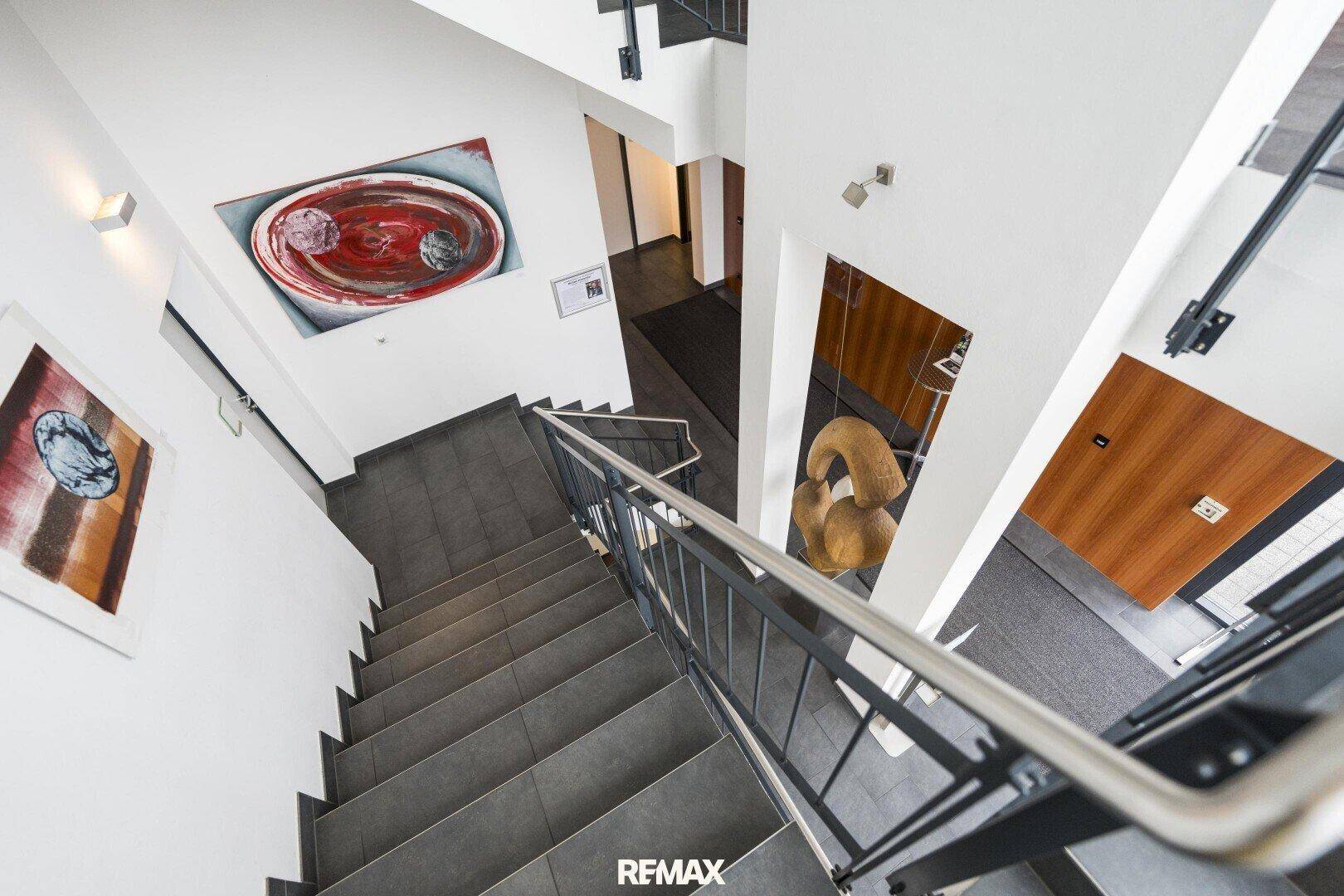 Blick von oben auf ein modernes Treppenhaus mit dunklen Fliesenstufen, Metallgeländer und Kunstwerken an den Wänden.