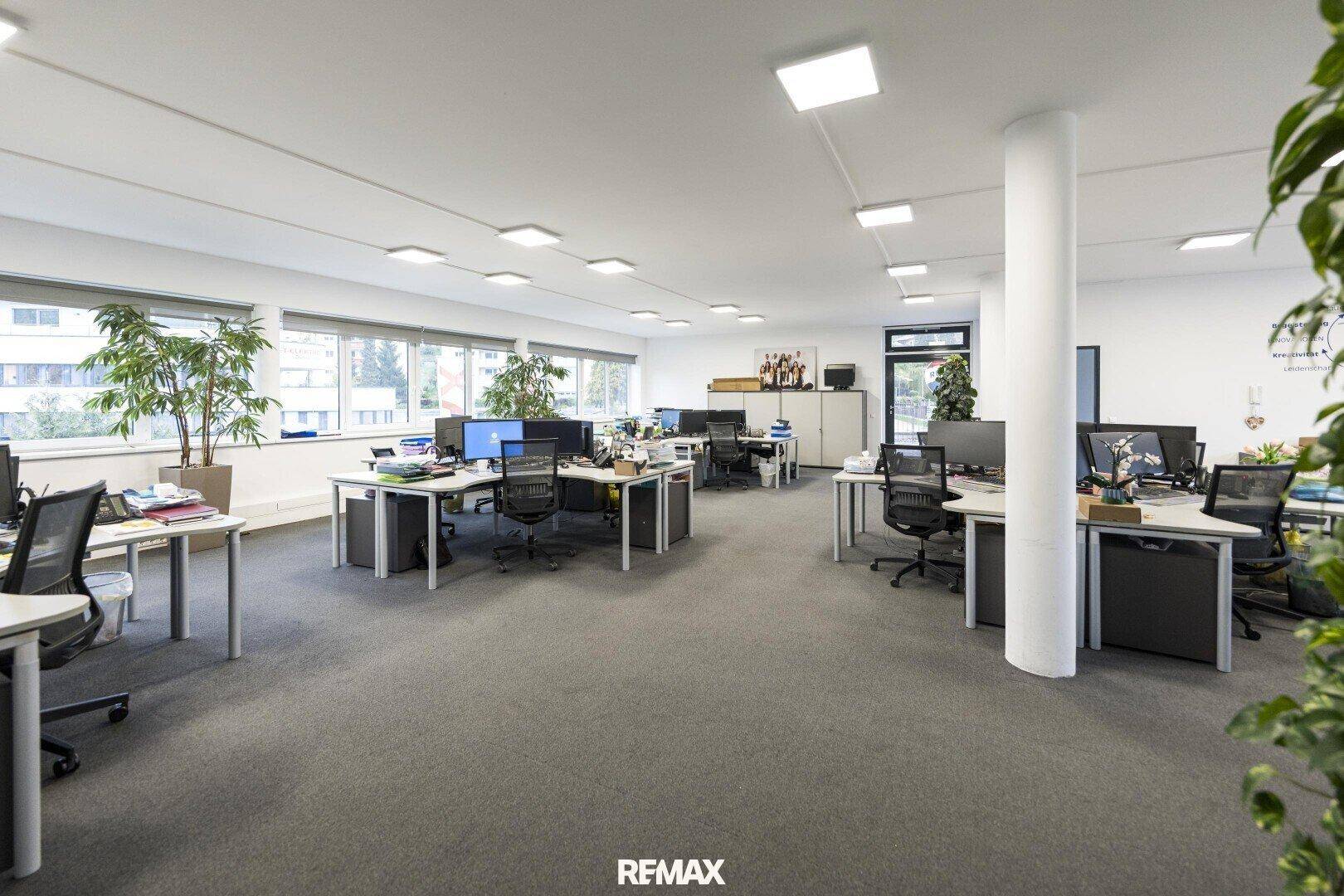 Weitläufiges Großraumbüro mit vielen Arbeitsplätzen, Teppichboden und Säulen, die den Raum strukturieren.