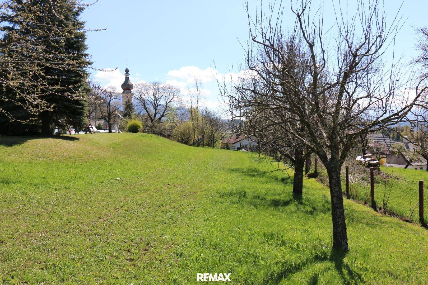 Weitläufiger grüner Gartenbereich mit Bäumen und einem Kirchturm, eingebettet in eine hügelige Landschaft.