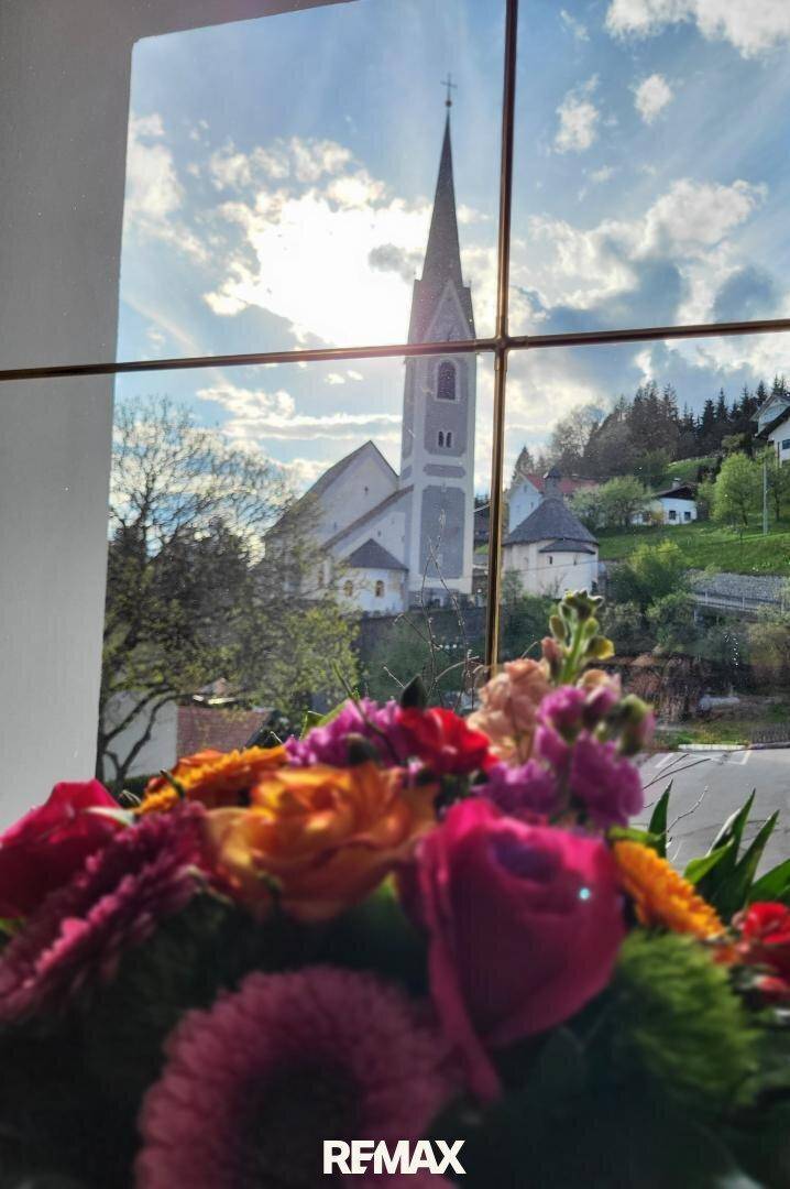 Blick aus dem Fenster auf eine malerische Kirche und die umliegende grüne Landschaft unter blauem Himmel.