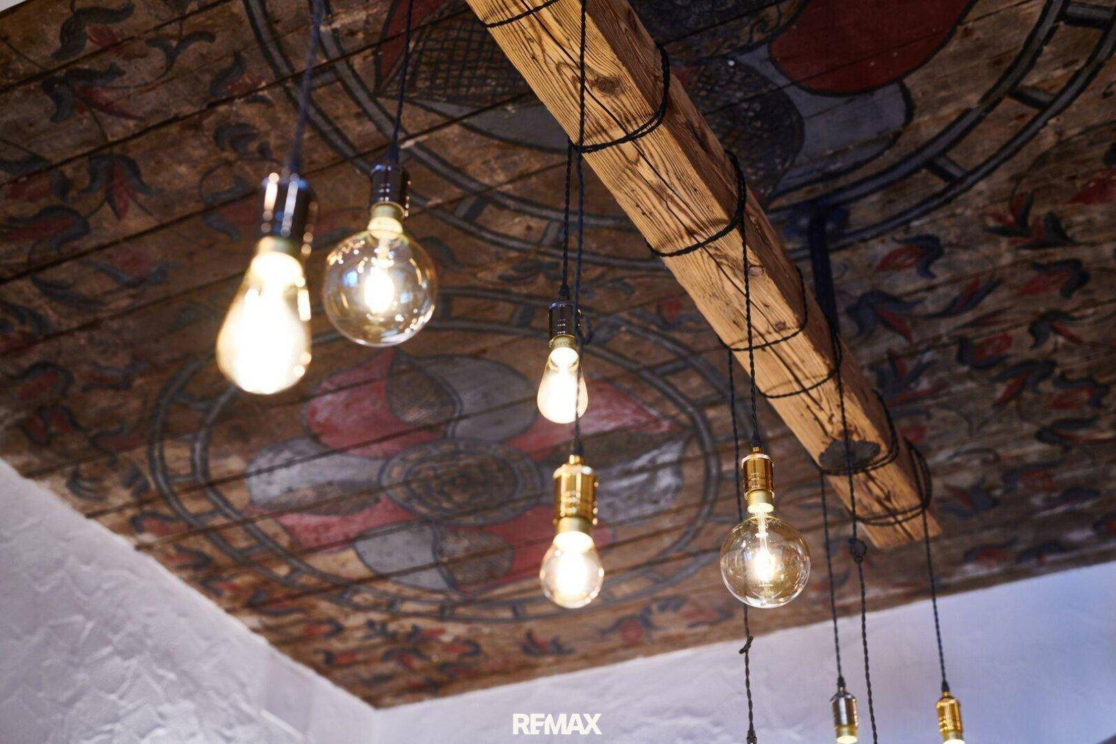 Historische Holzdecke mit kunstvollen Malereien und sichtbaren Balken, beleuchtet von dekorativen Glühbirnen.