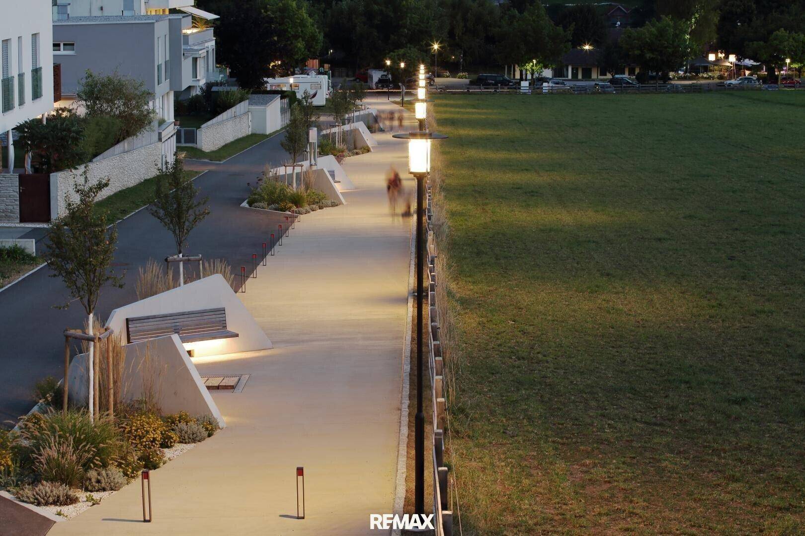 Beleuchtete Promenade entlang einer Grünfläche mit Bänken und moderner Beleuchtung bei Dämmerung.