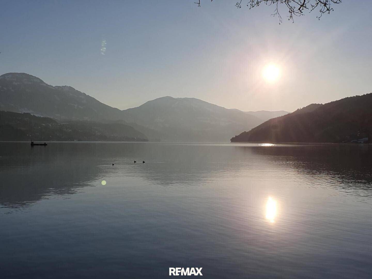 Malerische Seelandschaft bei Sonnenaufgang oder -untergang mit Bergen im Hintergrund und Sonnenreflexionen.