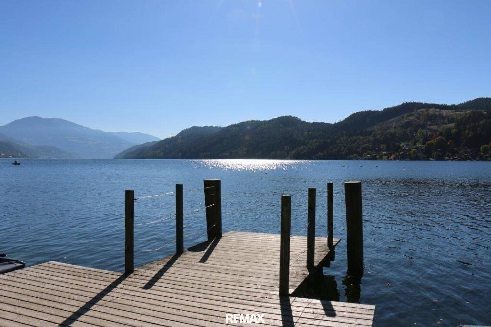 Hölzerner Steg ragt in einen ruhigen See, umgeben von Bergen unter einem strahlend blauen Himmel.