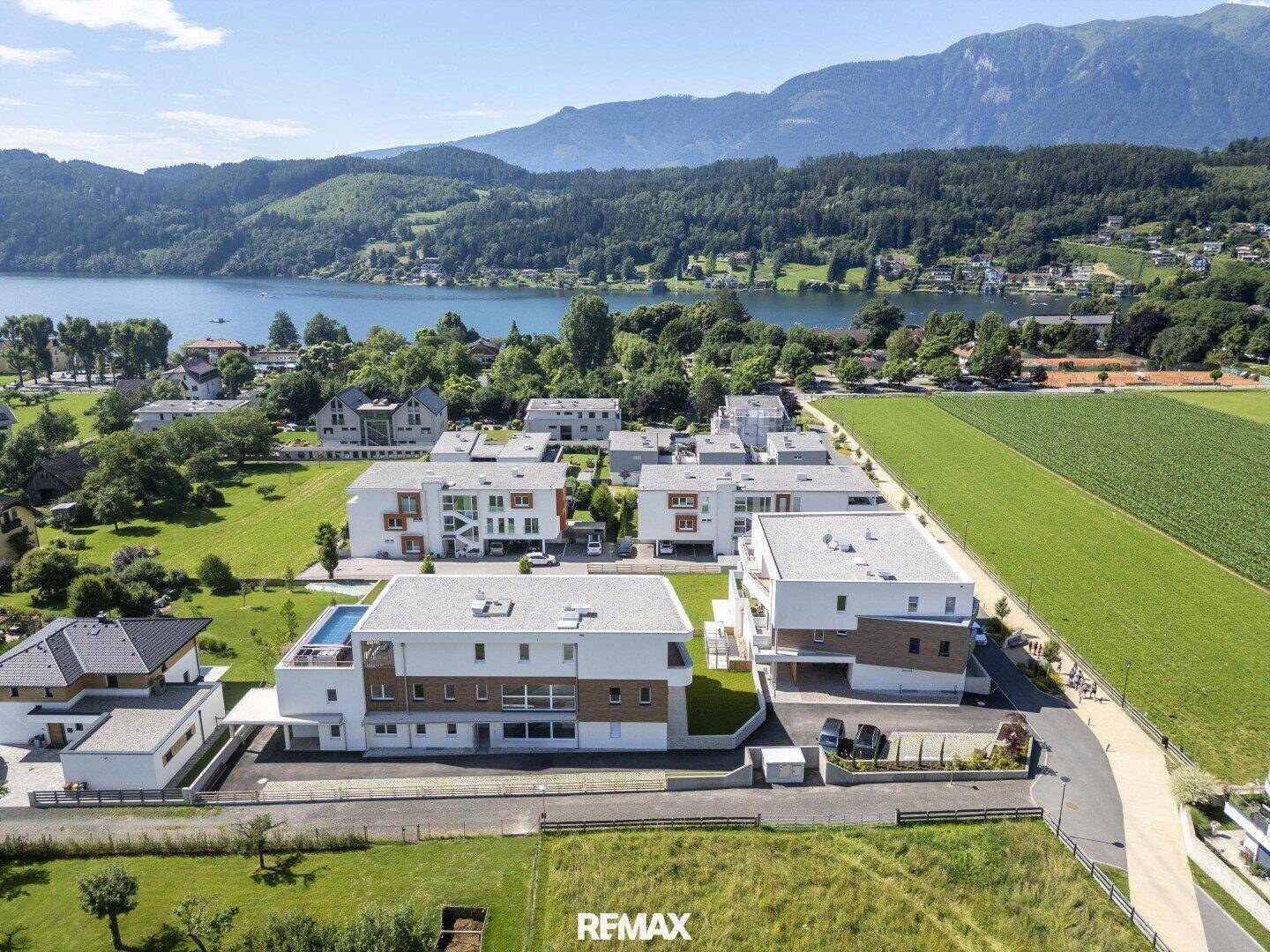 Luftaufnahme einer Wohnanlage mit modernen Gebäuden, teilweise mit Pools, nahe einem See und Bergen.
