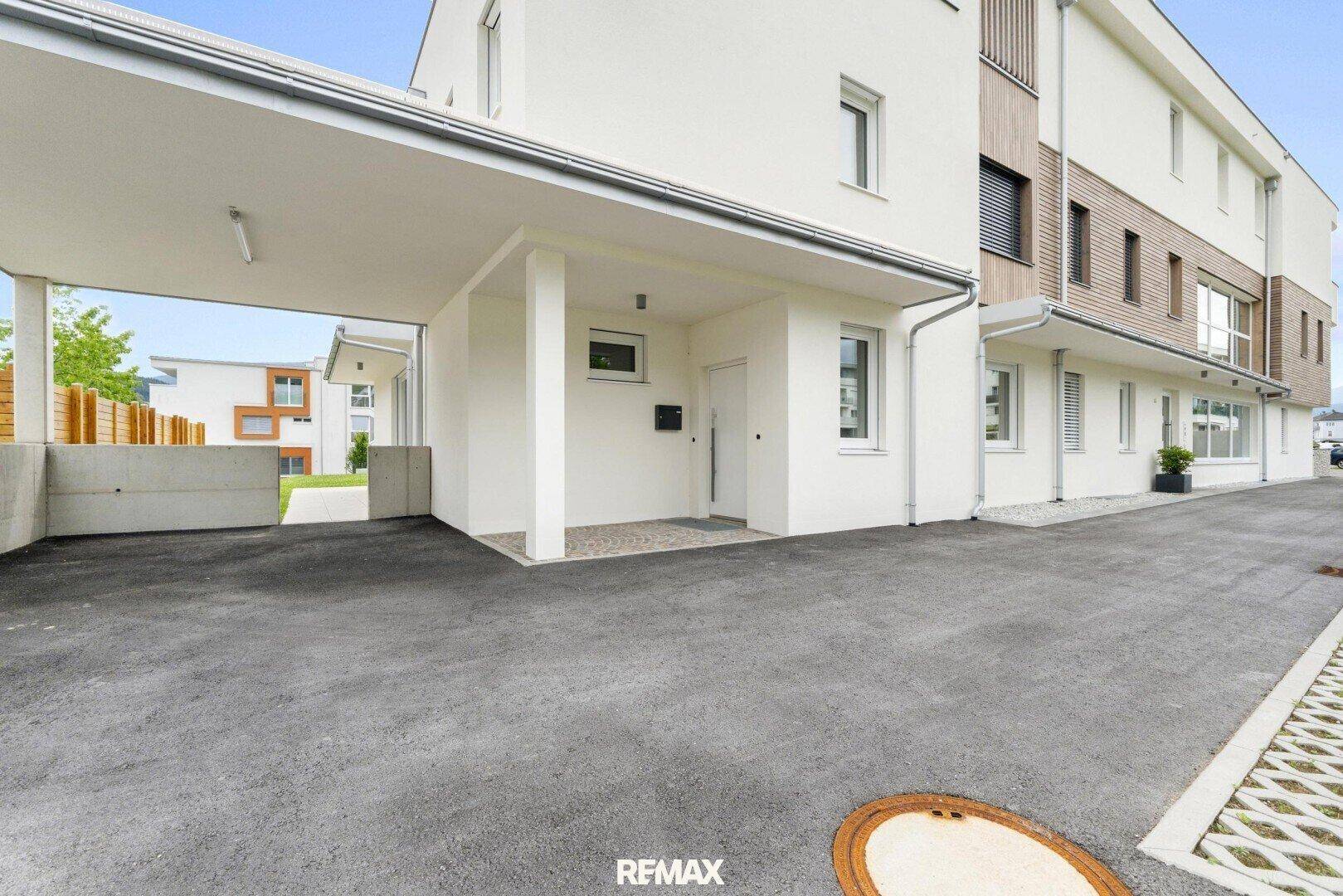 Eingangsbereich eines modernen Gebäudes mit überdachtem Carport und gepflastertem Zugang.