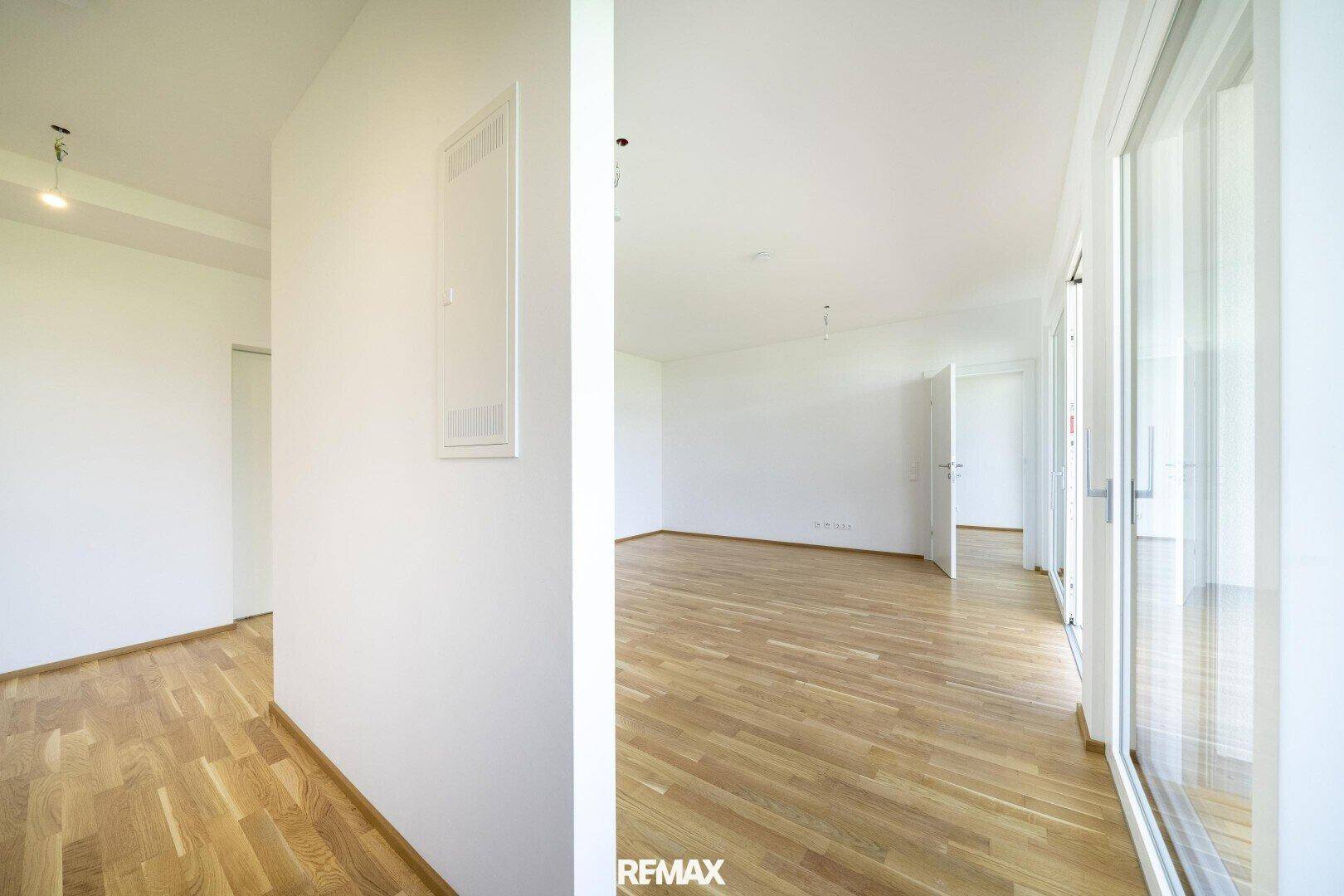 Heller, leerer Raum mit hellem Holzfußboden und großen Fenstertüren, die nach draußen führen.