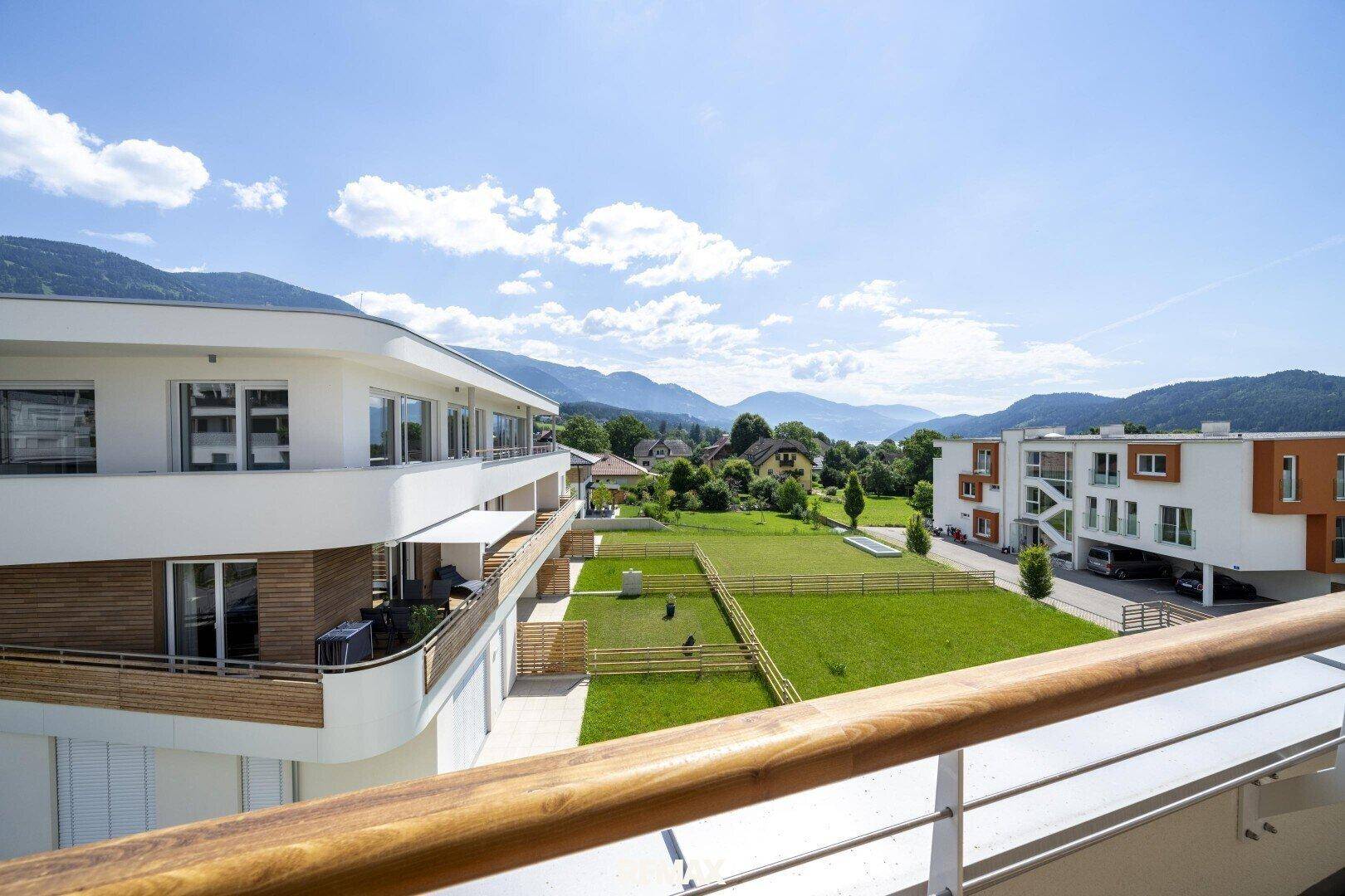 Ausblick von einem Balkon auf moderne Gebäude, grüne Gärten und eine Berglandschaft.