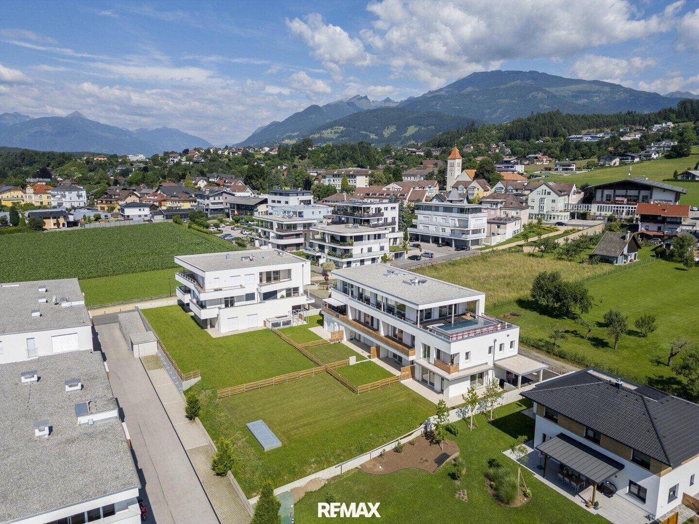 Luftaufnahme einer modernen Wohnsiedlung eingebettet in eine grüne Hügellandschaft mit Bergblick.