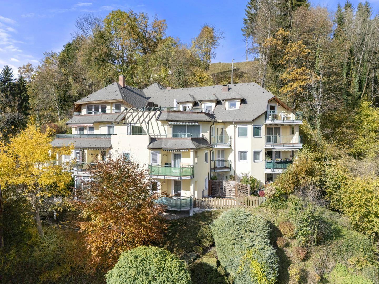 Mehrstöckiges Wohngebäude in Hanglage, umgeben von üppiger Herbstvegetation und Bäumen.