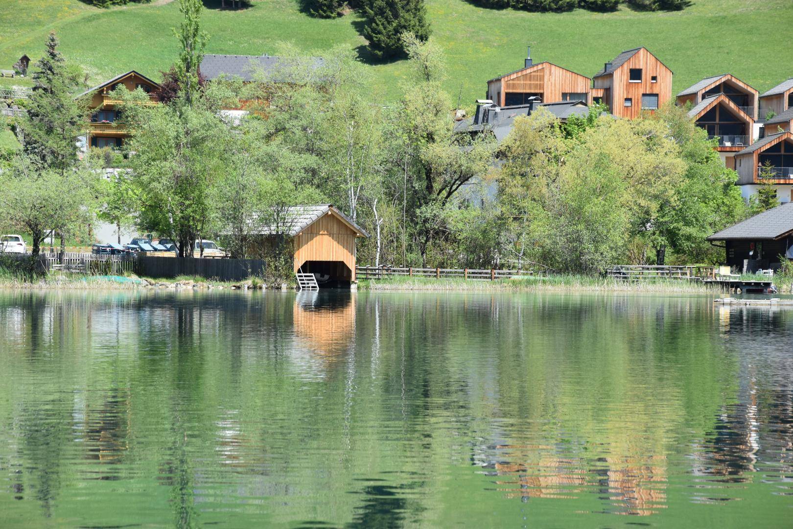 Idyllische Szene mit einem Holzbootshaus am Seeufer, umgeben von grünen Hügeln und Häusern.