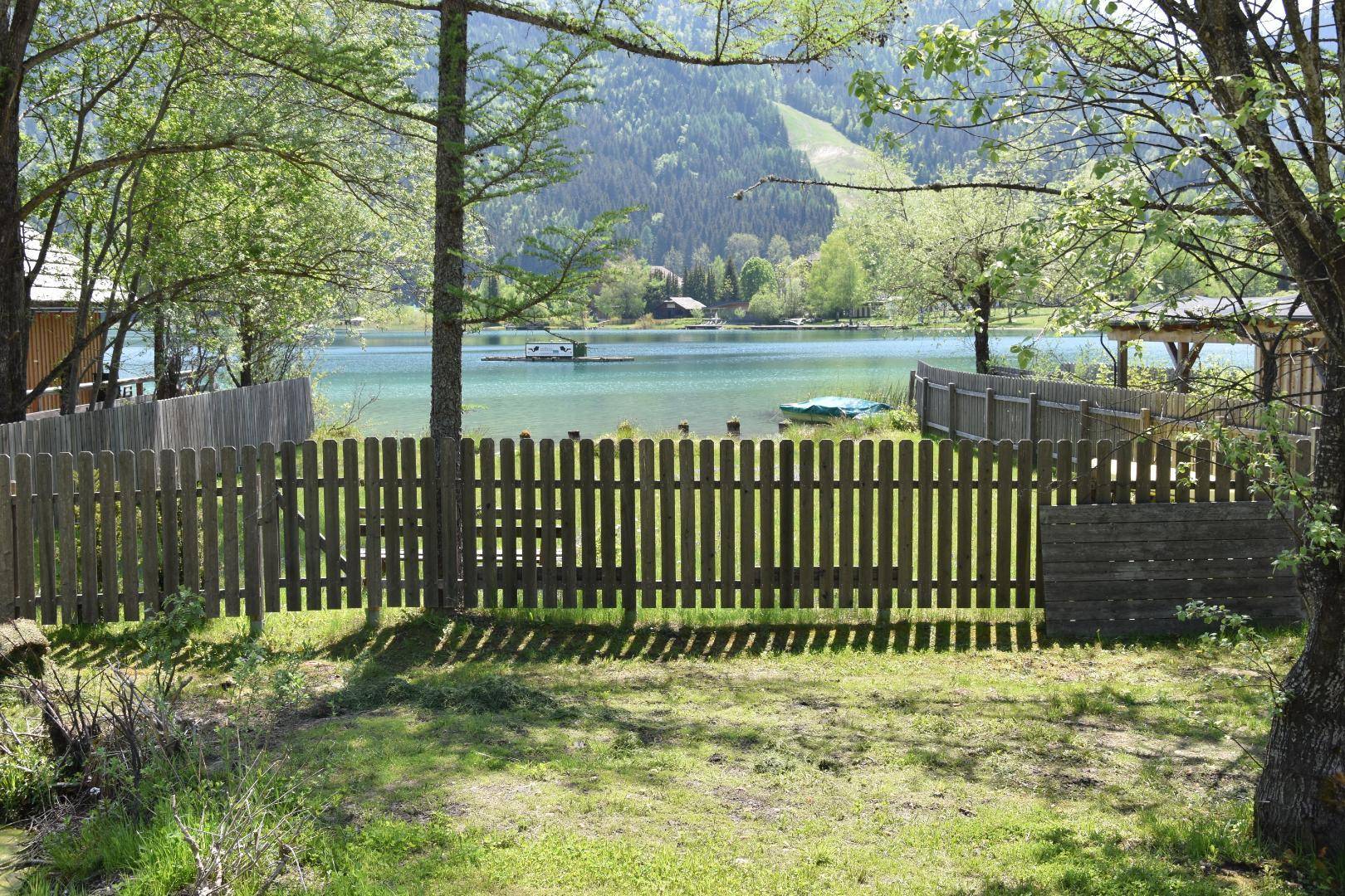 Blick über einen Holzzaun auf den ruhigen See und die bewaldeten Berge im Hintergrund.