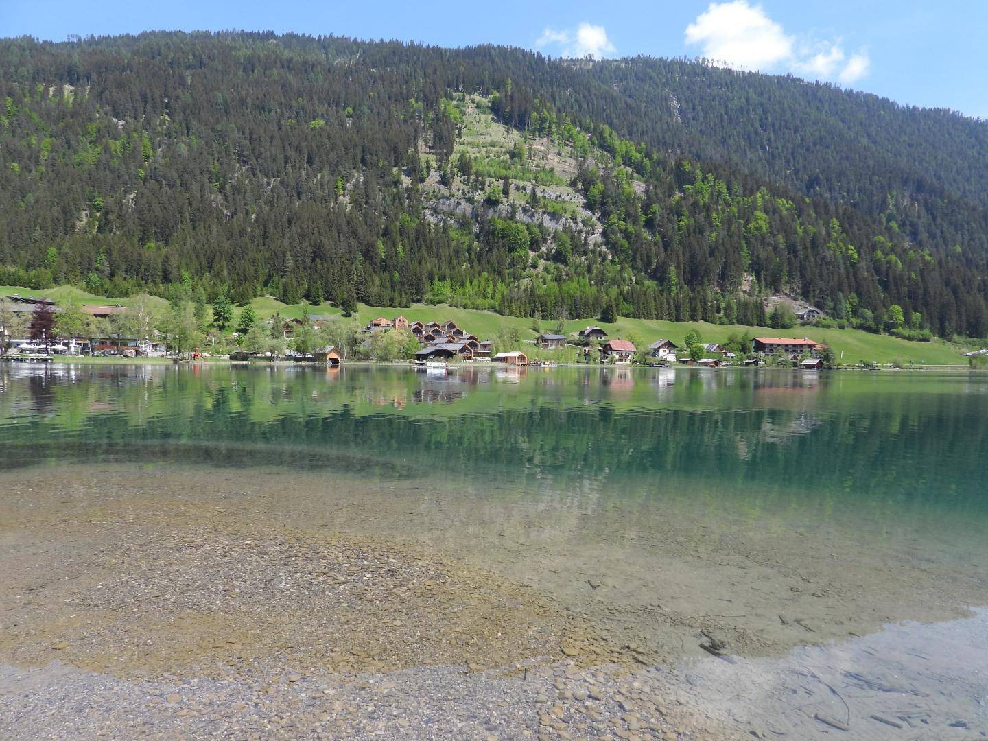 Klarer Blick über das türkisfarbene Wasser des Sees auf die bewaldeten Berghänge und die Uferbebauung.
