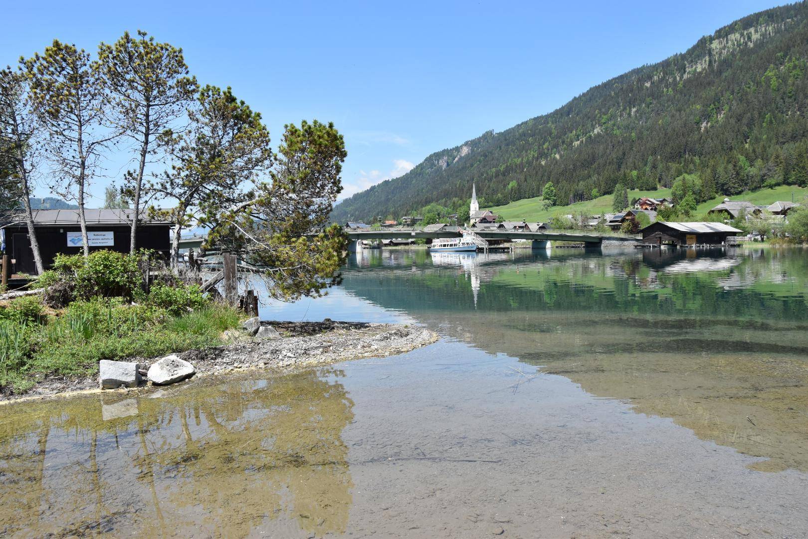 Weitblick über den See mit einer Brücke, einem Boot und der malerischen Uferlandschaft im Hintergrund.