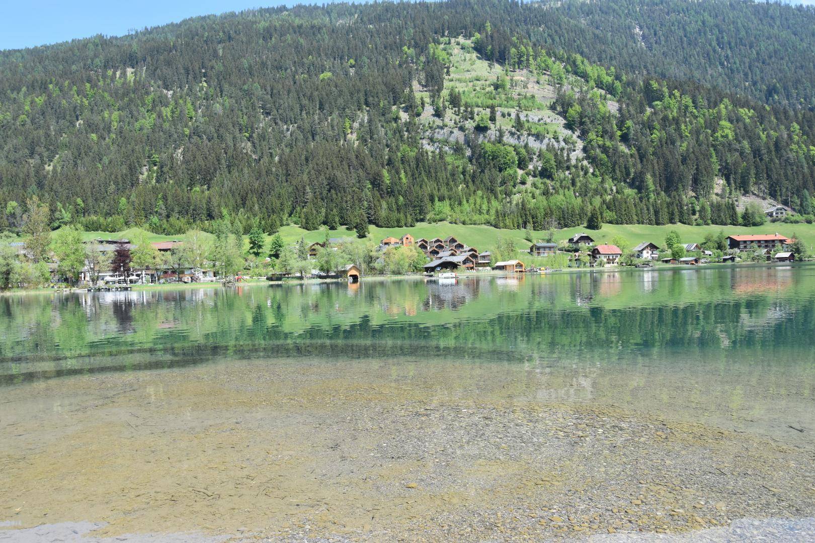 Malerischer Blick über den klaren See auf die gegenüberliegende Uferbebauung und bewaldete Berge.
