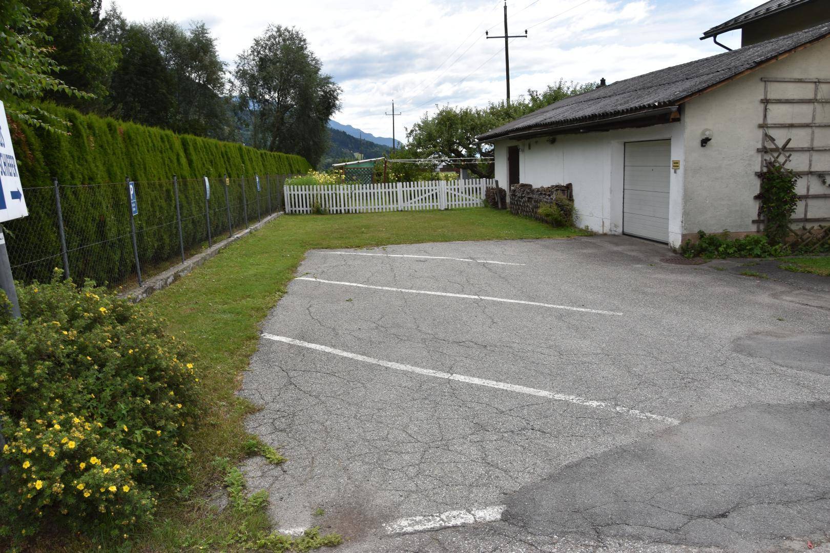 Außenbereich mit asphaltiertem Parkplatz und einem Garagengebäude, umgeben von Hecken und einem weißen Zaun.
