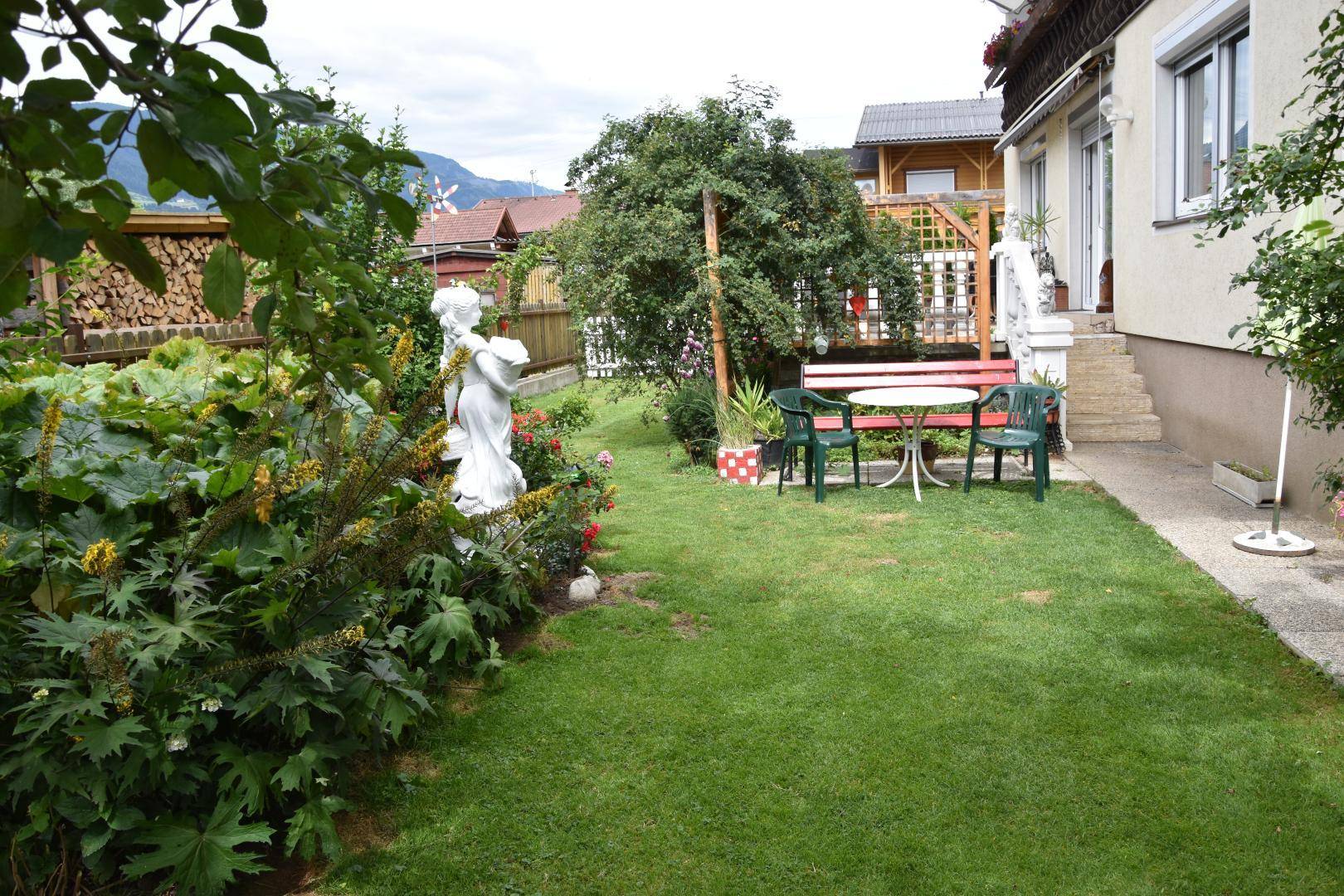 Großer, grüner Garten mit Rasenfläche, Blumenbeeten und einer Sitzgruppe vor dem Haus, umgeben von Natur.