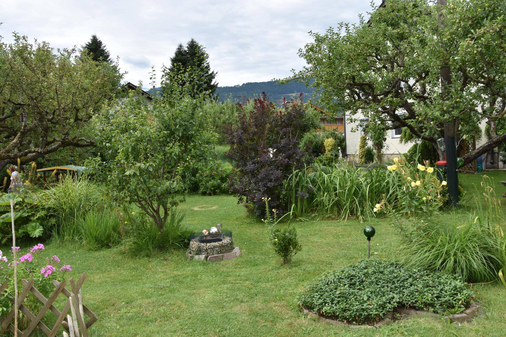 Weitläufiger Garten mit verschiedenen Pflanzen, Bäumen und einem kleinen Zierbrunnen im Vordergrund, umgeben von Natur.