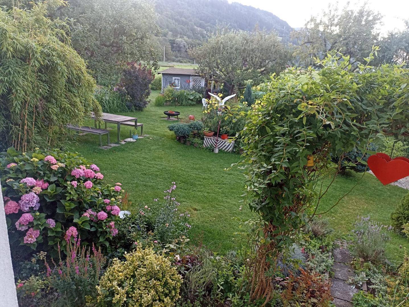 Weitläufiger Garten mit verschiedenen Pflanzen, Bäumen und einem Sitzbereich im Hintergrund, umgeben von Natur.