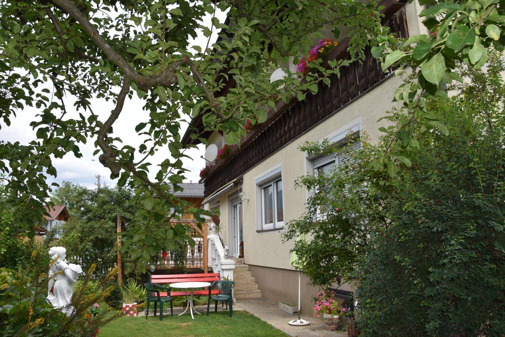Gepflegter Garten mit Rasen, Blumen und einer roten Sitzgruppe vor dem Haus mit Balkon und grüner Umgebung.