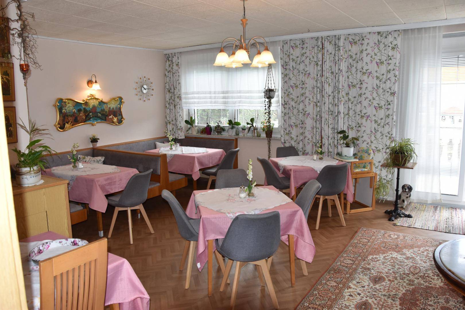 Heller Speisesaal mit mehreren Tischen, Sitzbänken und floralen Vorhängen, ideal für Gäste und Mahlzeiten.