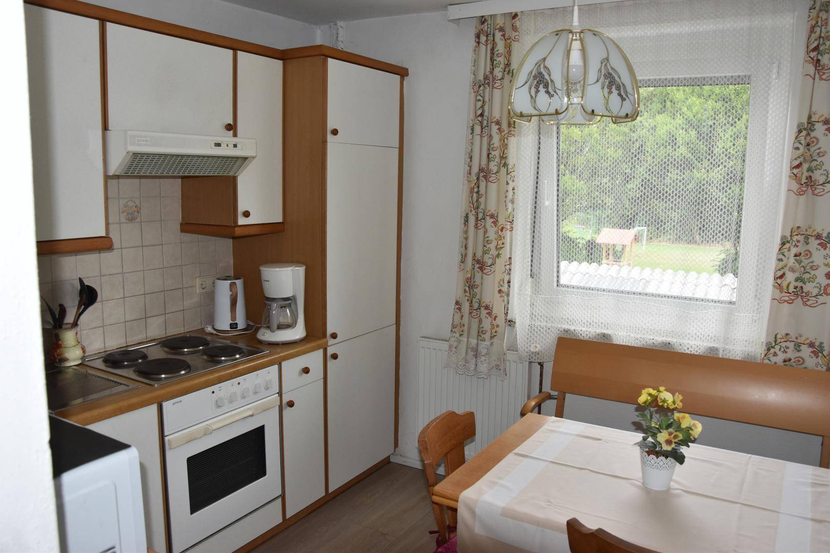 Funktionale Küche mit weißen Schränken, Elektroherd und einem Esstisch am Fenster mit Blick ins Grüne.