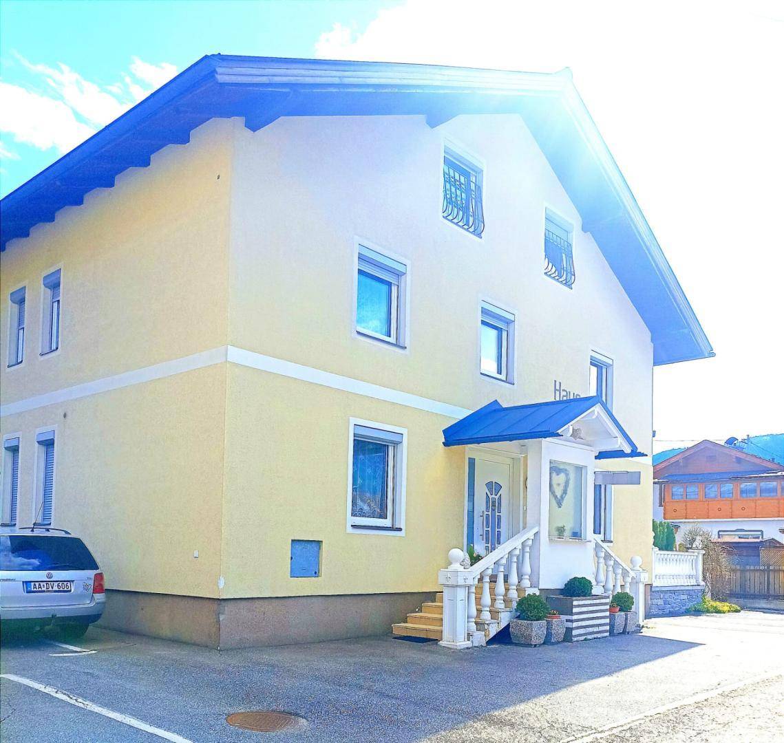 Gelbe Hausfassade mit blauem Dach und Eingangsbereich unter strahlendem Sonnenschein.