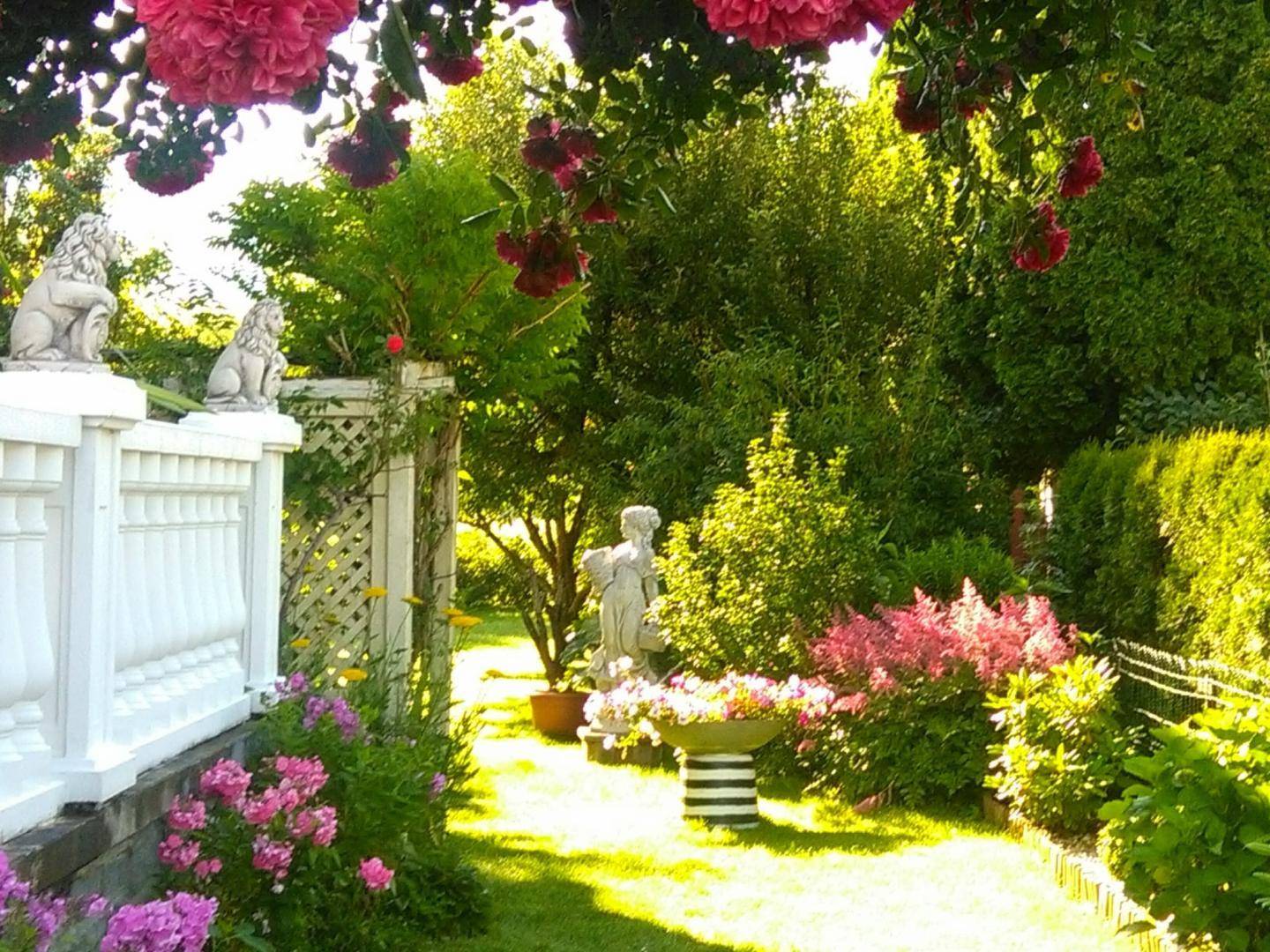 Sonnige Gartenecke mit Balustrade, Blumenbeeten und dekorativen Statuen.