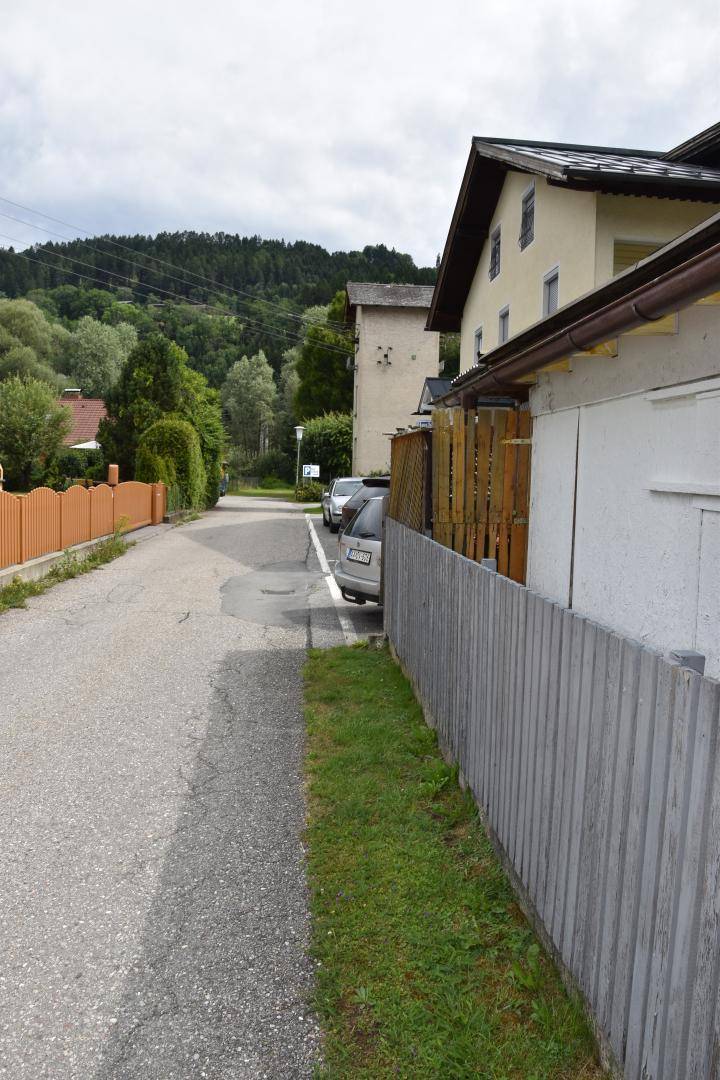 Ruhige Wohnstraße mit angrenzendem Haus und grüner Hügellandschaft im Hintergrund.