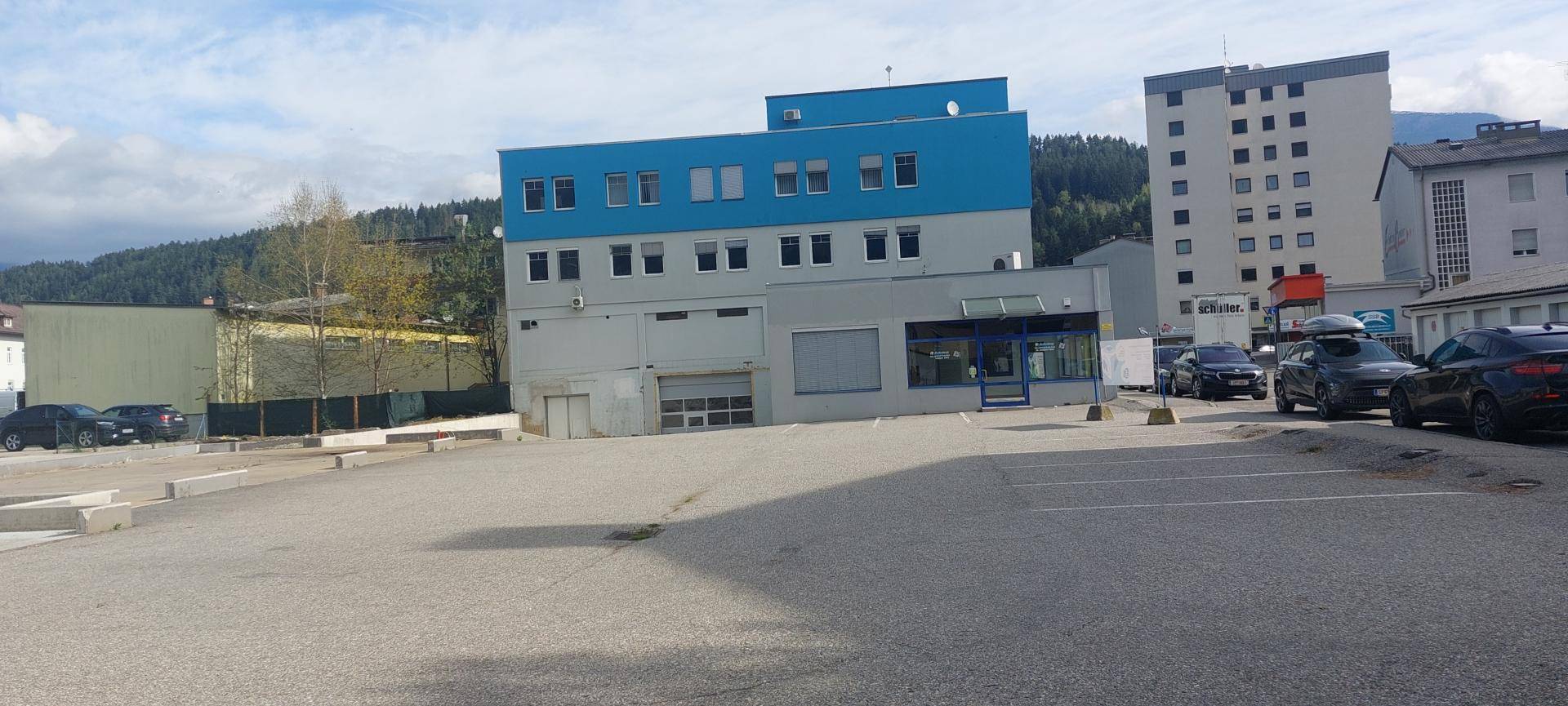 Gebäude mit blauer und grauer Fassade, umgeben von einem Parkplatz mit einigen Autos und Bergen im Hintergrund.