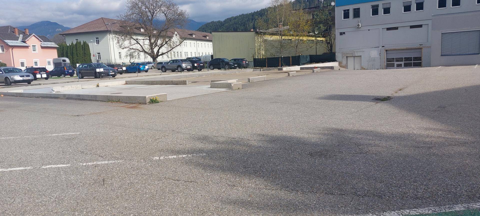 Großer asphaltierter Parkplatz mit Blick auf weitere Gebäude und eine bergige Landschaft im Hintergrund.