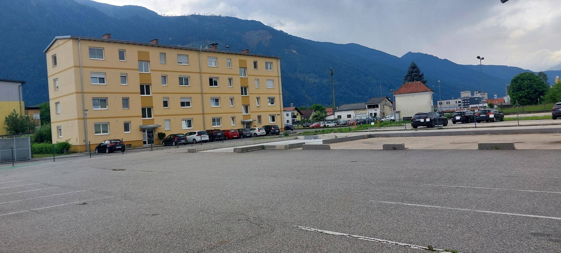 Weitläufiger Parkplatz mit einem gelben Wohngebäude und Bergen im Hintergrund unter bewölktem Himmel.