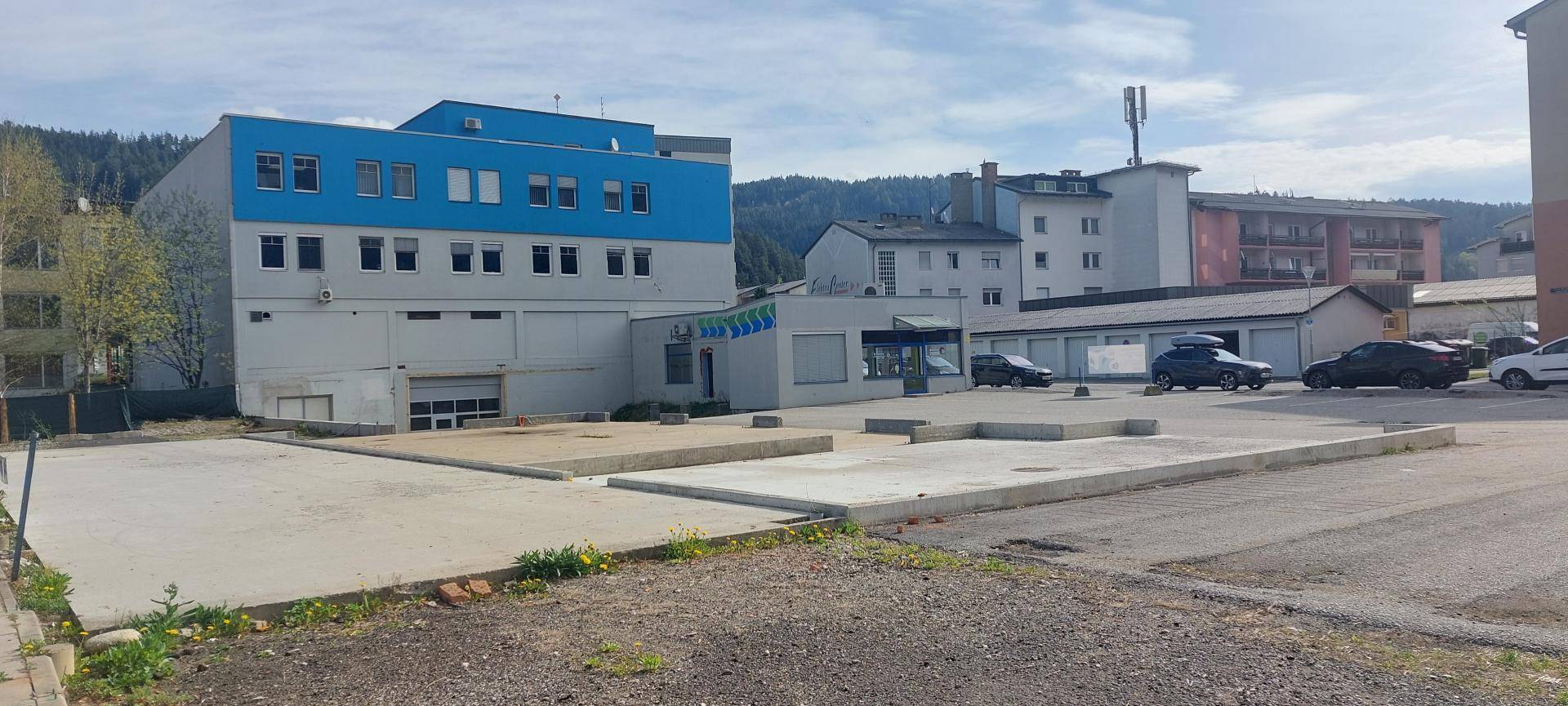 Ansicht des Gebäudes mit blauer und grauer Fassade, umgeben von einem leeren Parkplatz und anderen Gebäuden.