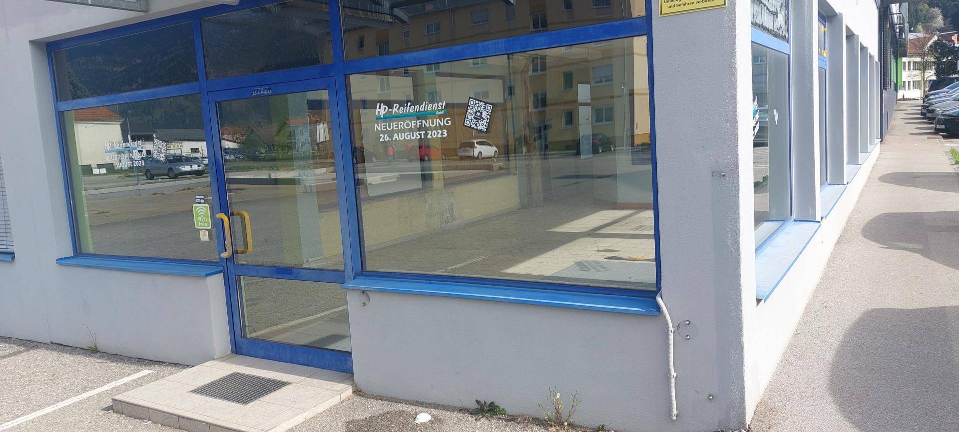 Eingangsbereich eines Geschäfts mit großen blauen Fensterrahmen und einer Glastür, die die Umgebung widerspiegelt.