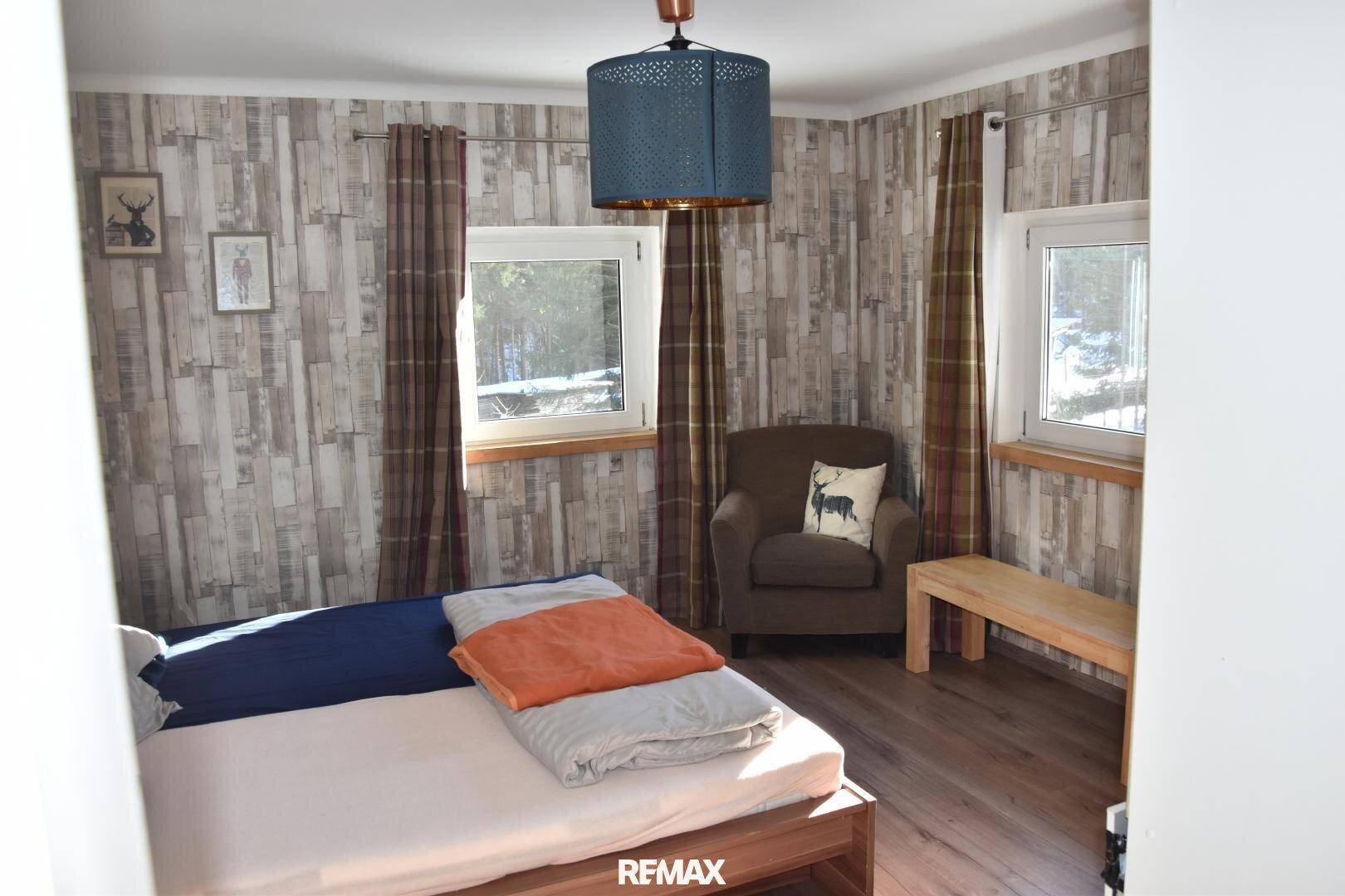 Gemütliches Schlafzimmer mit Holzboden, einem Sessel und zwei Fenstern für viel Tageslicht.