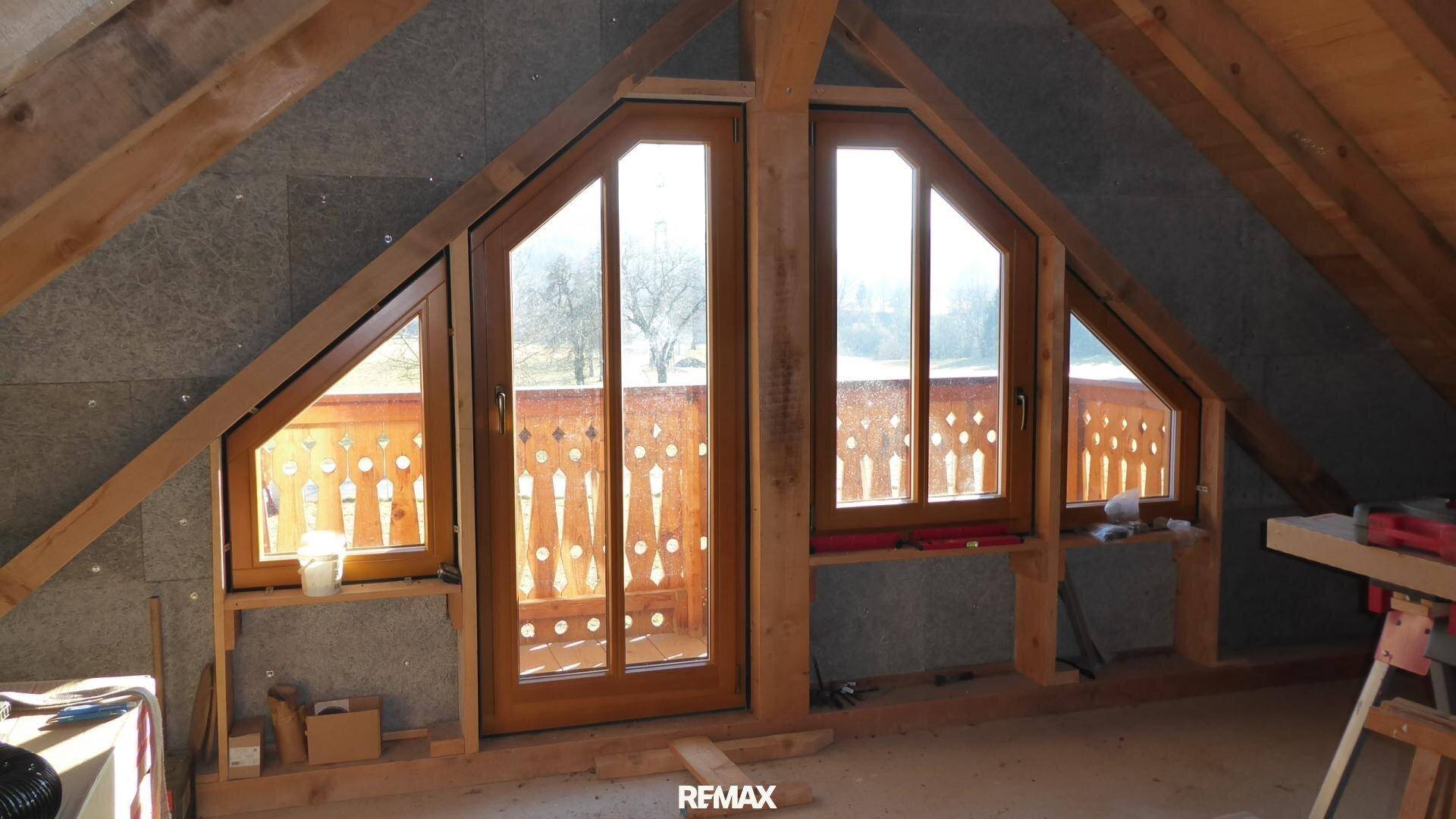 Dachgeschoss im Rohbau mit schrägen Holzdecken und großen Fenstern zum Balkon.