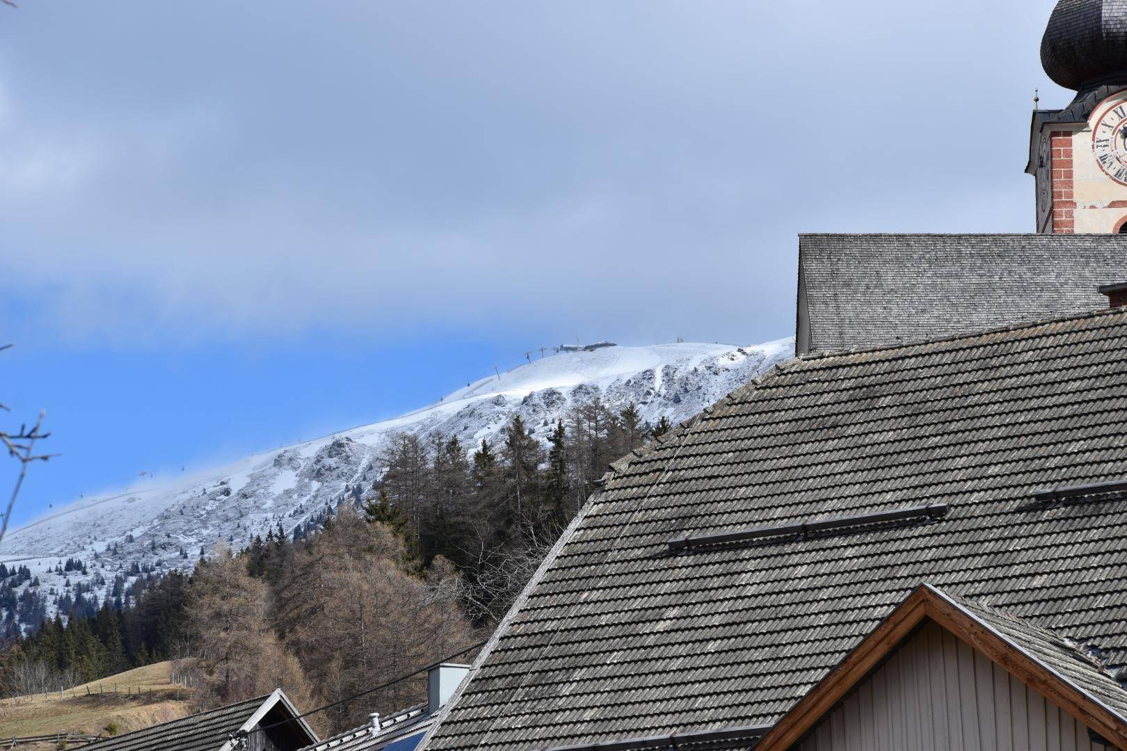 Blick über Dächer auf eine schneebedeckte Berglandschaft mit Kirchturm.