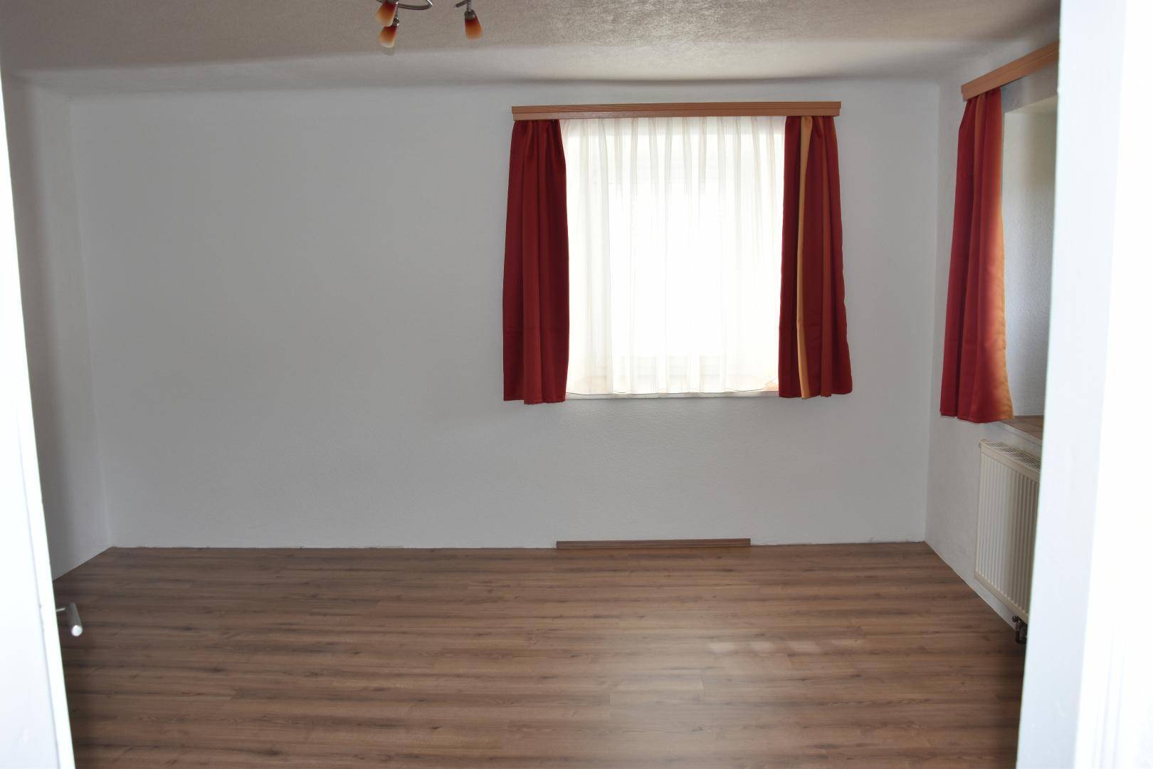 Heller Raum mit Laminatboden und roten Vorhängen an einem großen Fenster.