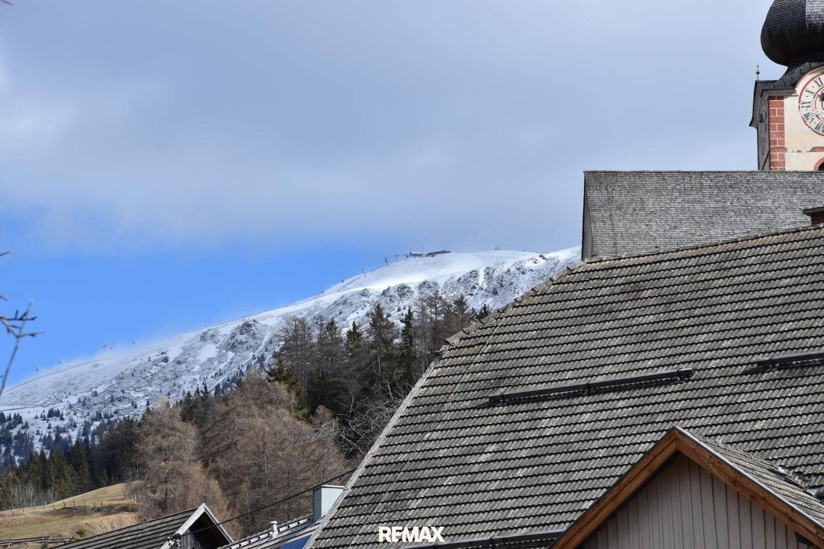 Panoramablick auf schneebedeckte Berge und traditionelle Dächer mit Kirchturm in der Ferne.