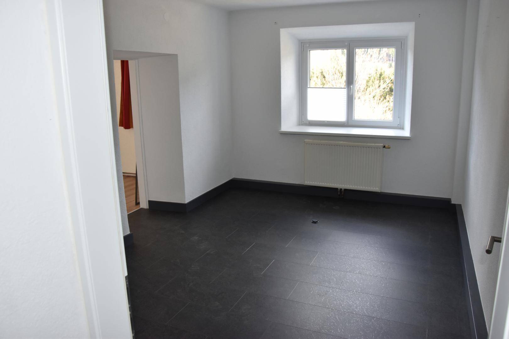 Leerer Raum mit dunklem Fliesenboden und einem Fenster, das Tageslicht hereinlässt.