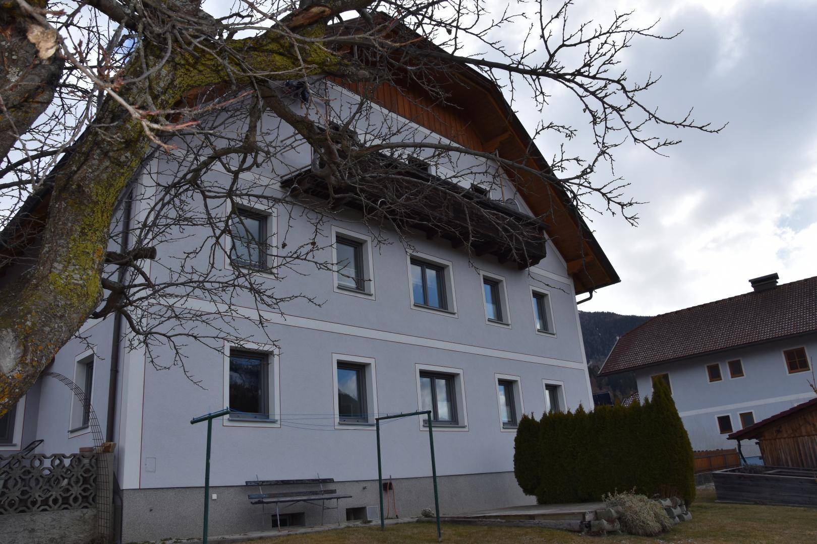 Seitenansicht des Hauses mit grauer Fassade, Fenstern und einem Baum im Vordergrund.