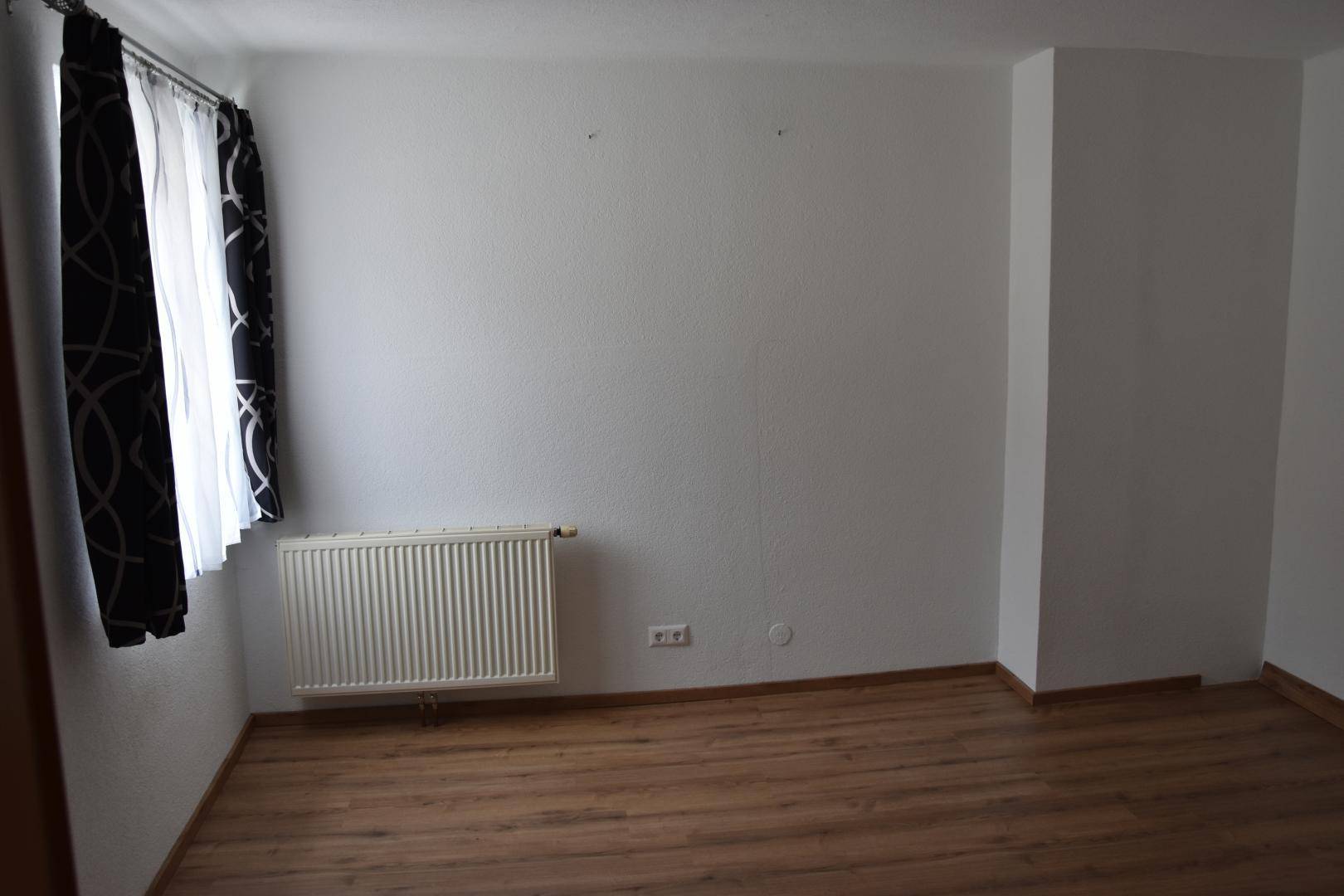 Leerer Raum mit weiß gestrichenen Wänden, Laminatboden und einem Heizkörper.