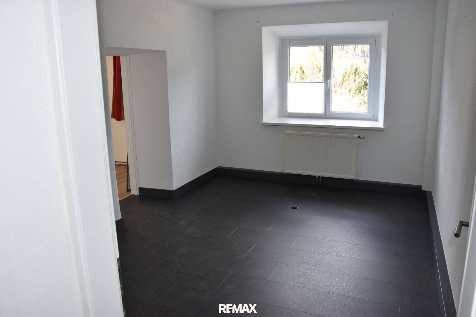 Leerer Raum mit dunklem Fliesenboden und einem Fenster, das Tageslicht hereinlässt.