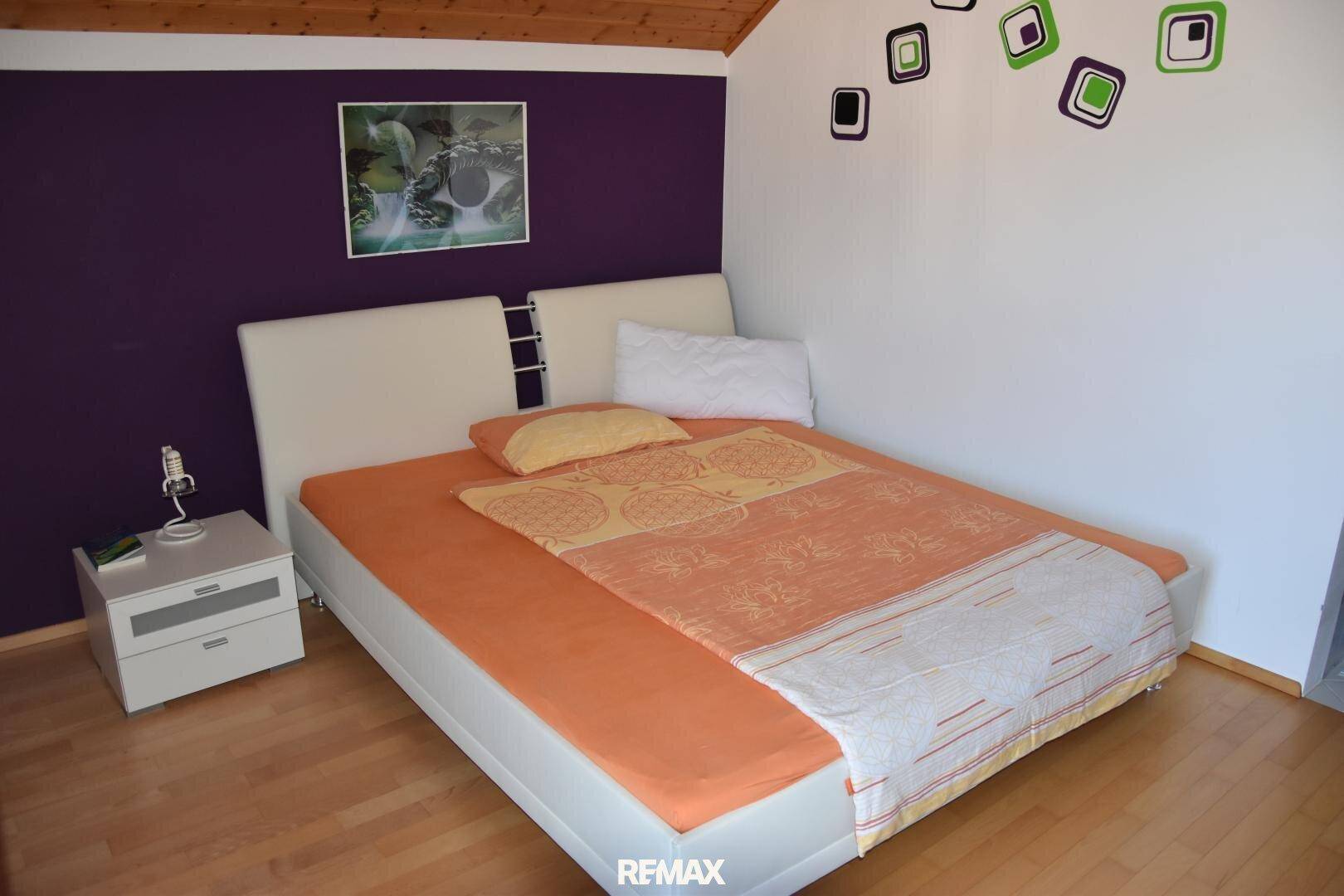 Schlafzimmer mit Doppelbett, lila Akzentwand und Holzfußboden, modern eingerichtet.