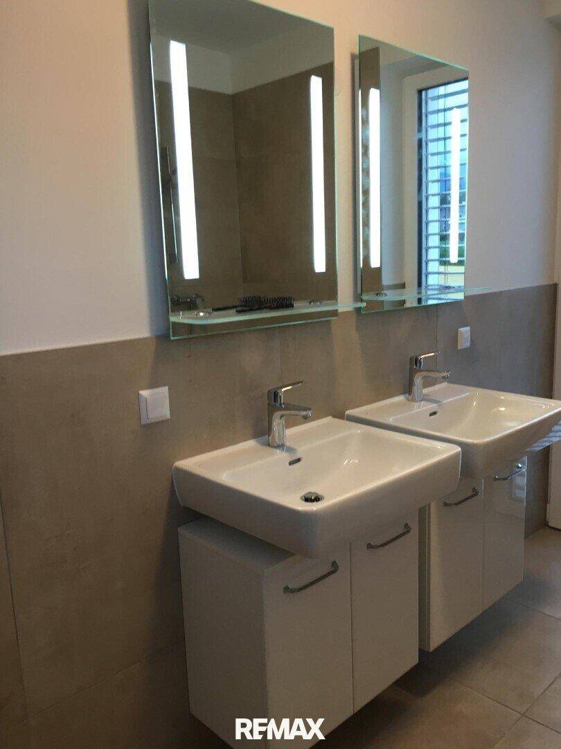 Modernes Badezimmer mit zwei Waschbecken, beleuchteten Spiegeln und hellen Fliesen.