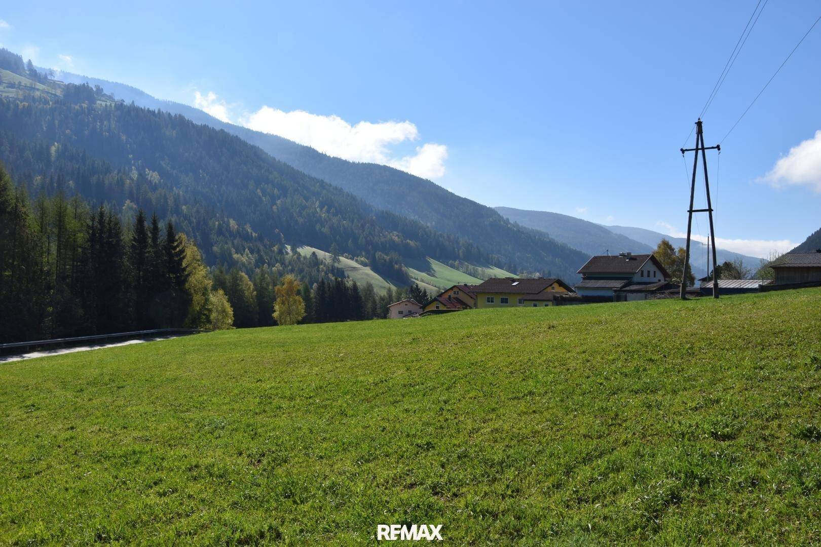 Weitläufiges grünes Feld mit Blick auf bewaldete Berge und einige Häuser in der Ferne unter blauem Himmel.