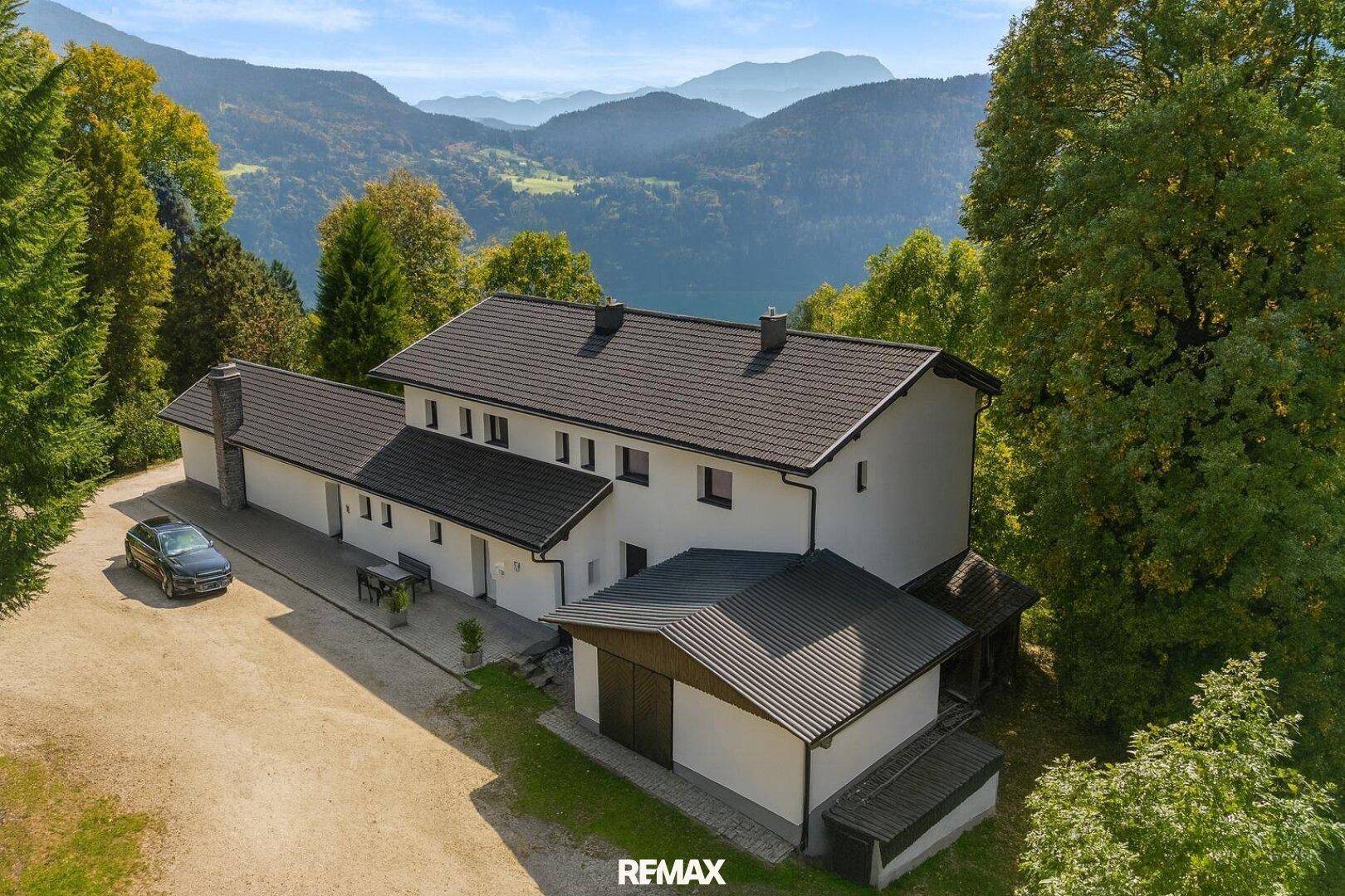 Luftaufnahme des Hauses mit großem Vorplatz, umgeben von Bäumen und Bergen im Hintergrund.