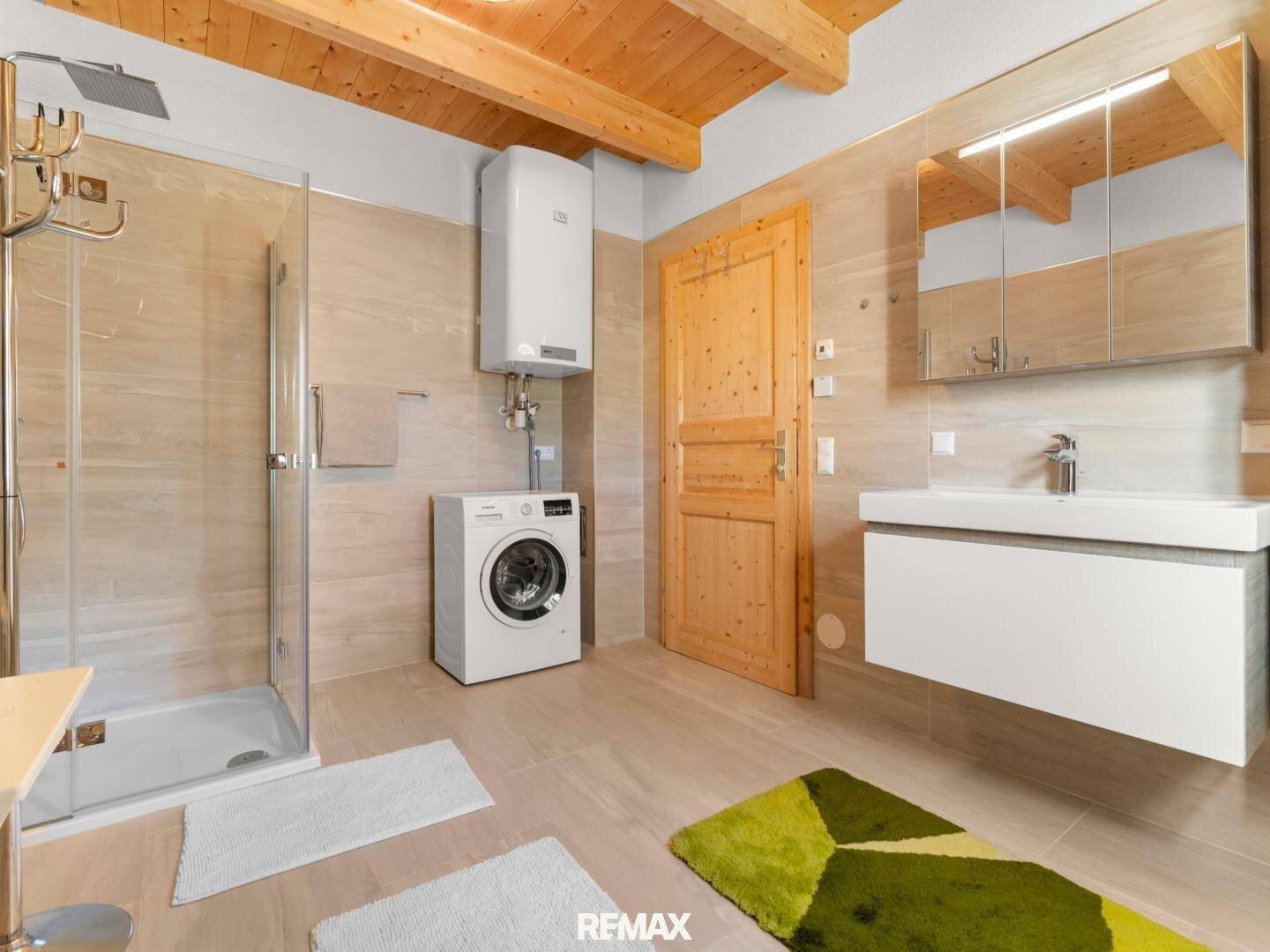 Modernes Badezimmer mit beigefarbenen Fliesen, Duschkabine, Waschtisch und Waschmaschine.