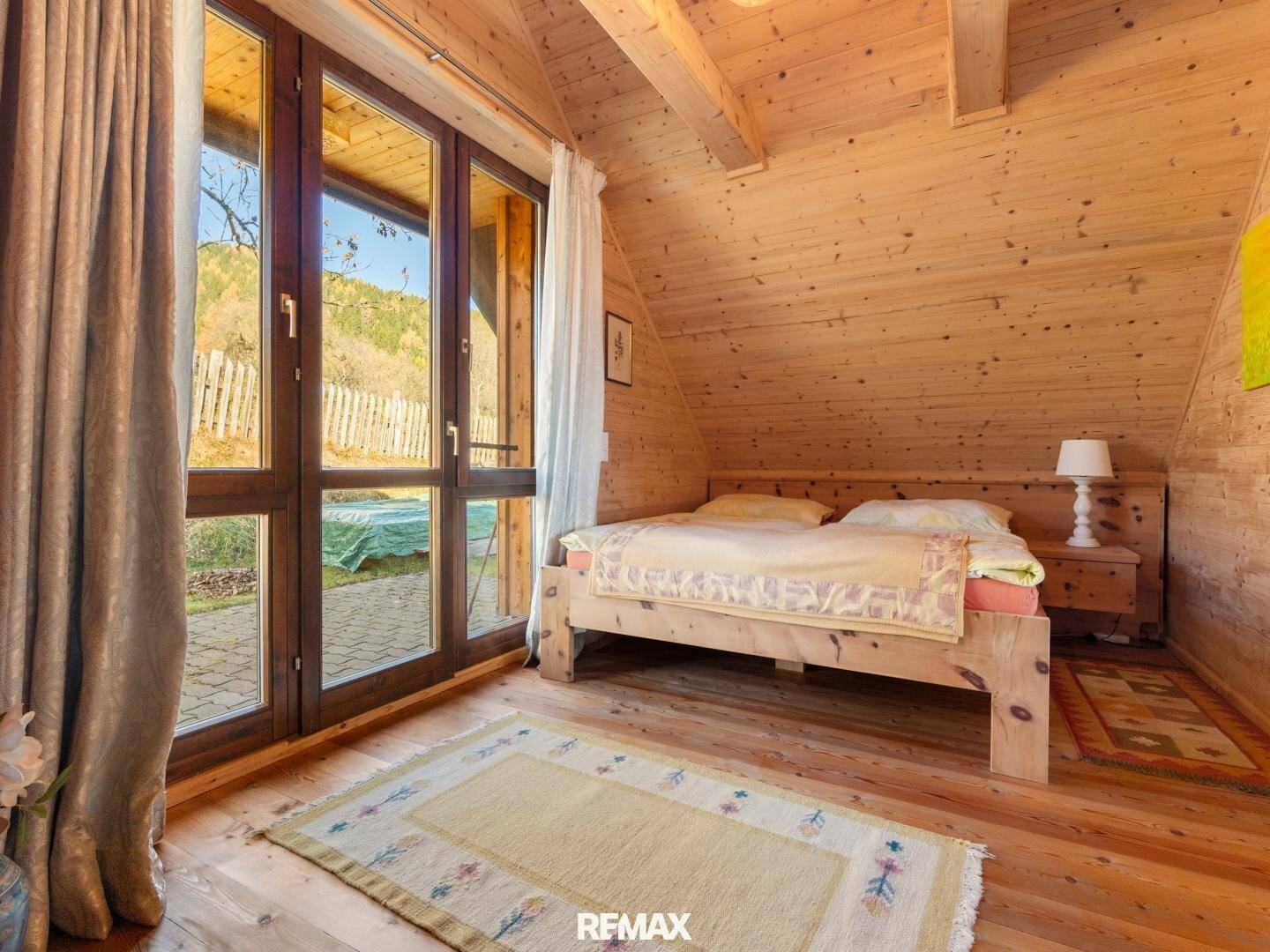 Gemütliches Schlafzimmer mit Holzwänden, Doppelbett und großen Fenstern mit Zugang nach draußen.