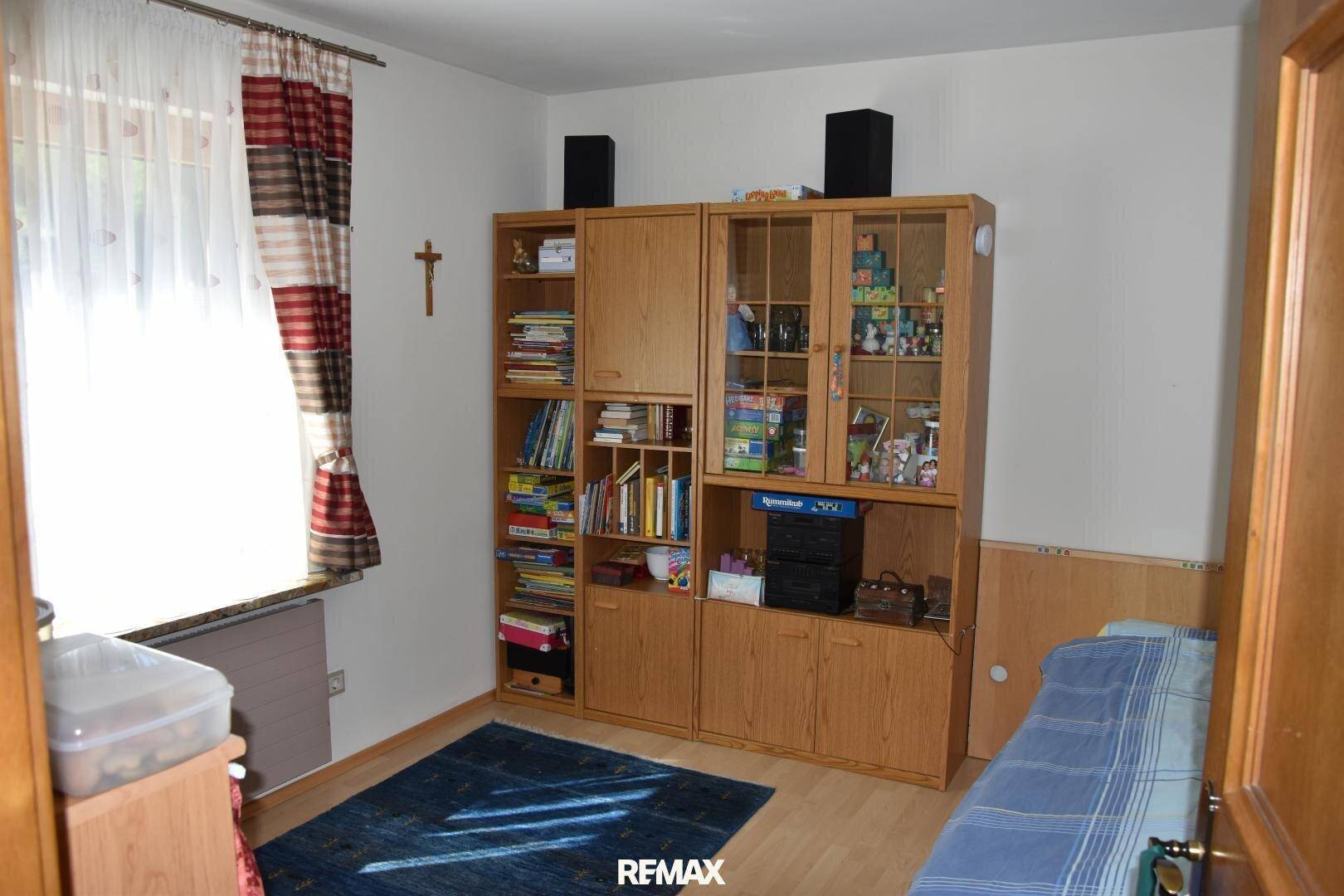 Helles Zimmer mit Einzelbett, Holzregal und einem Fenster, das viel Tageslicht hereinlässt.